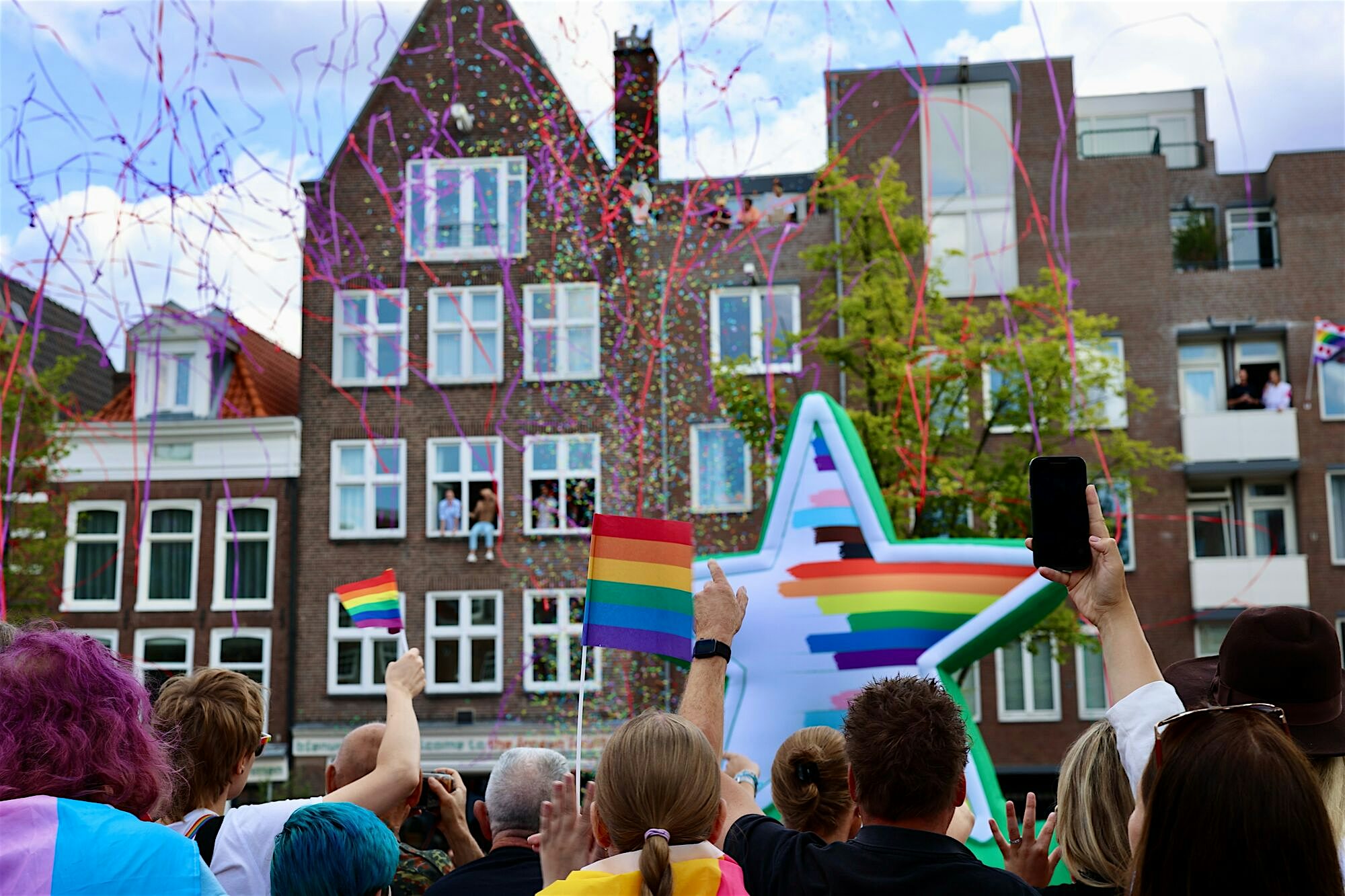 Crowd gathered along a canal raising rainbow flags as colourful confetti falls around a large star‑shaped float with rainbow stripes
