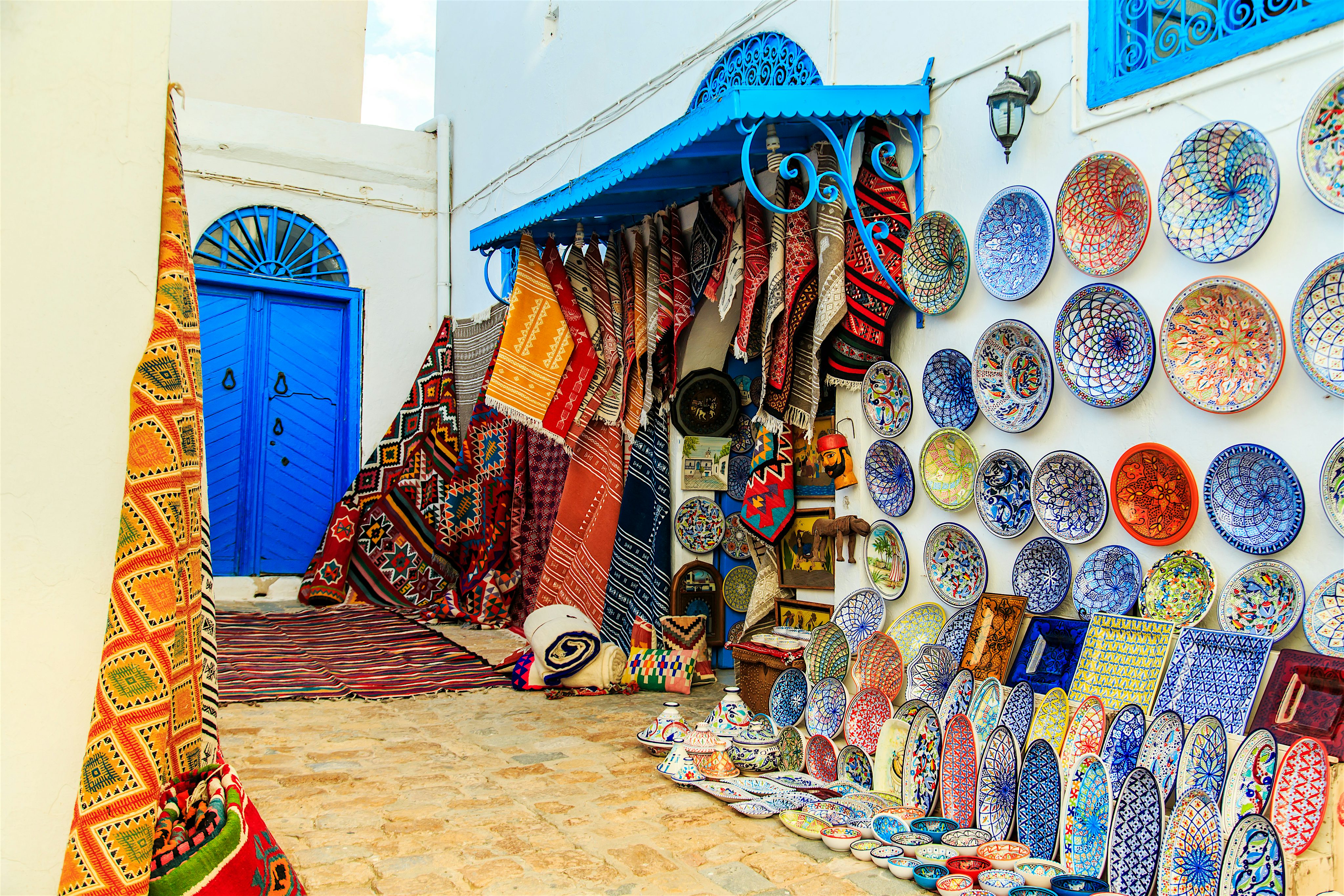 A narrow street market with colourful ceramics displayed on a wall and patterned rugs hanging beside a bright blue door.