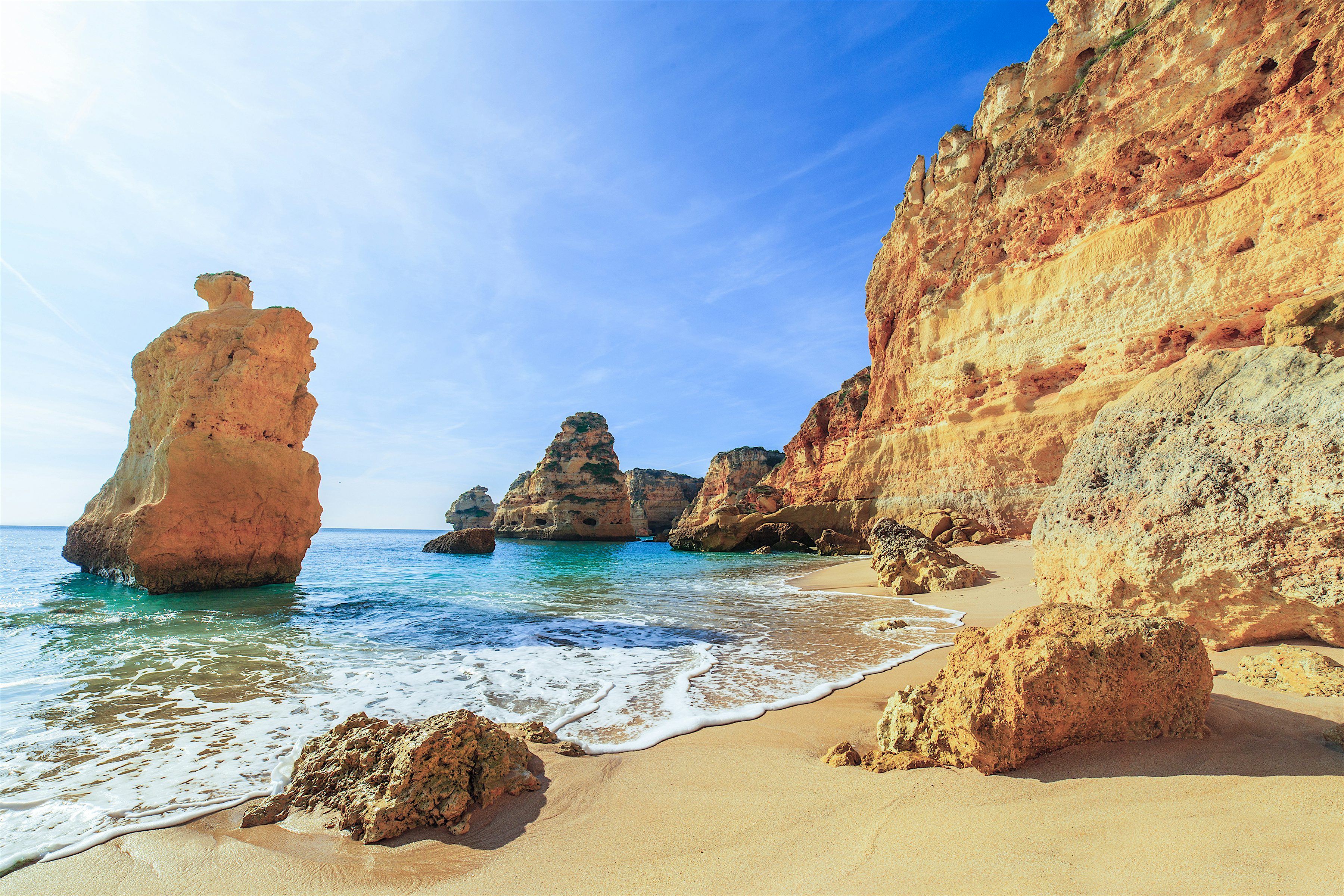 Golden sandstone cliffs and rock formations rise above a sandy beach with gentle waves