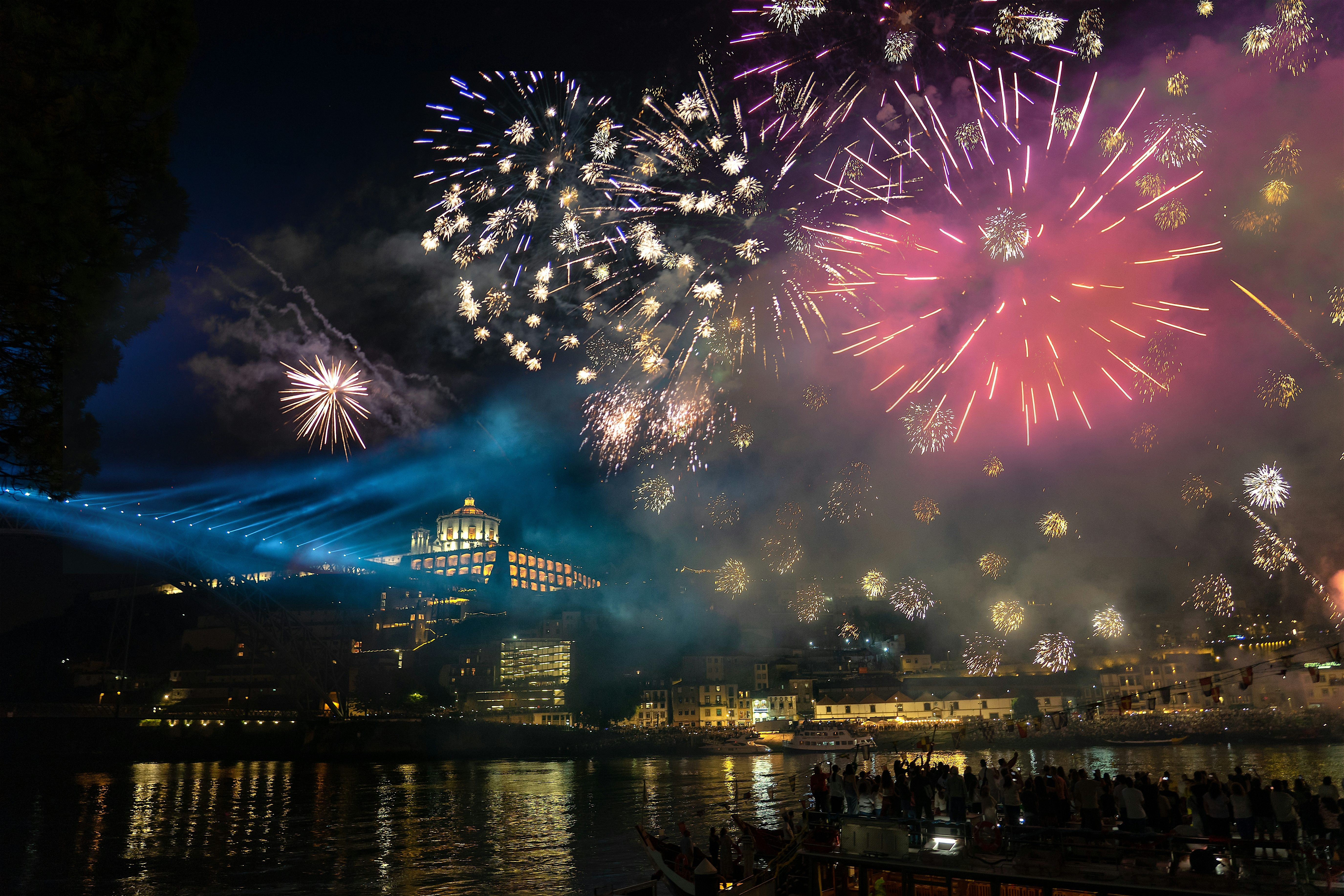 Fireworks in the night sky above a city, with bursts of colour reflecting on the water below, historic buildings lit along the shoreline, and a crowd gathered at the waterfront in the foreground.