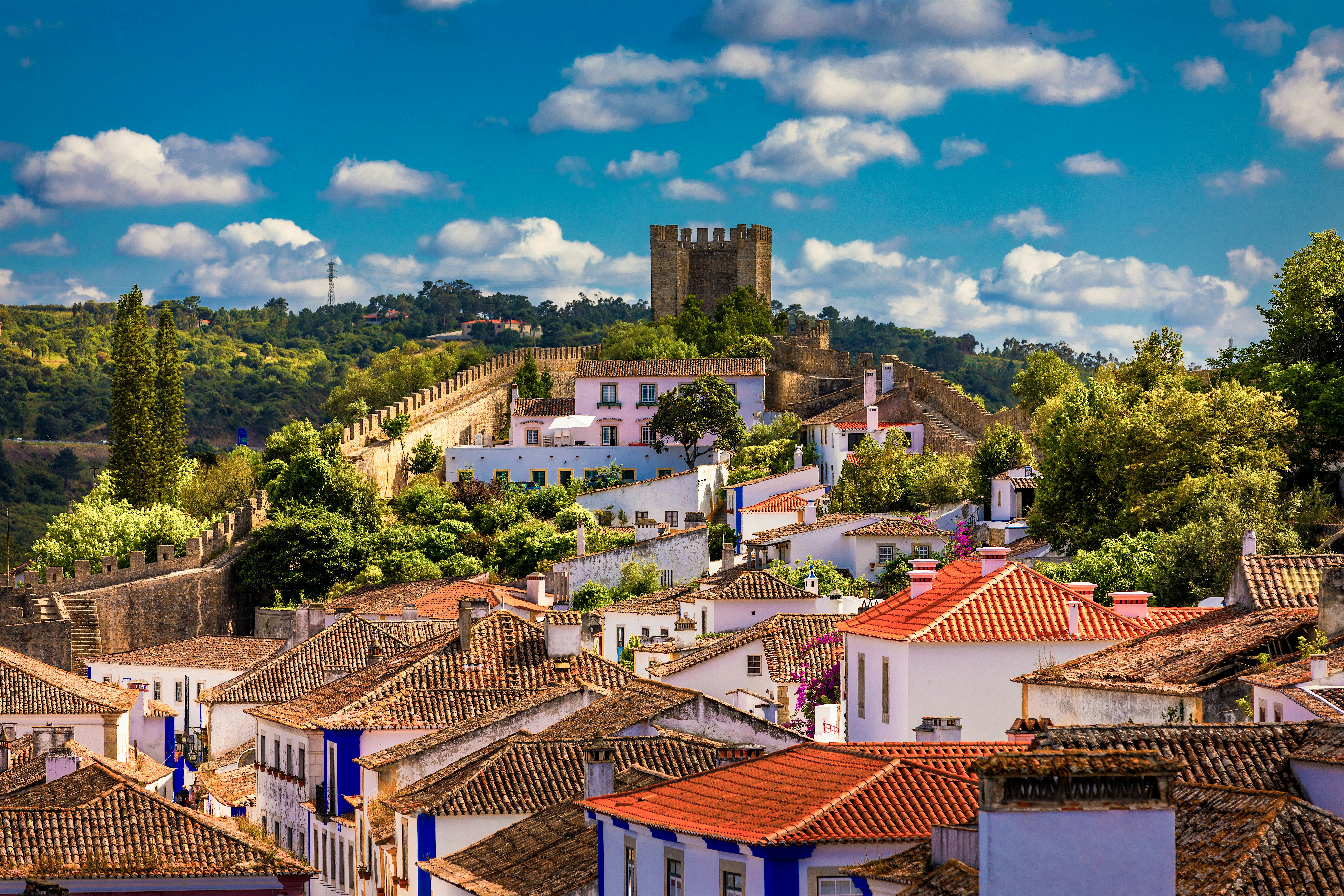 Whitewashed hillside town with terracotta rooftops and narrow streets, rising towards a stone castle and fortified walls set among green trees, with rolling countryside.