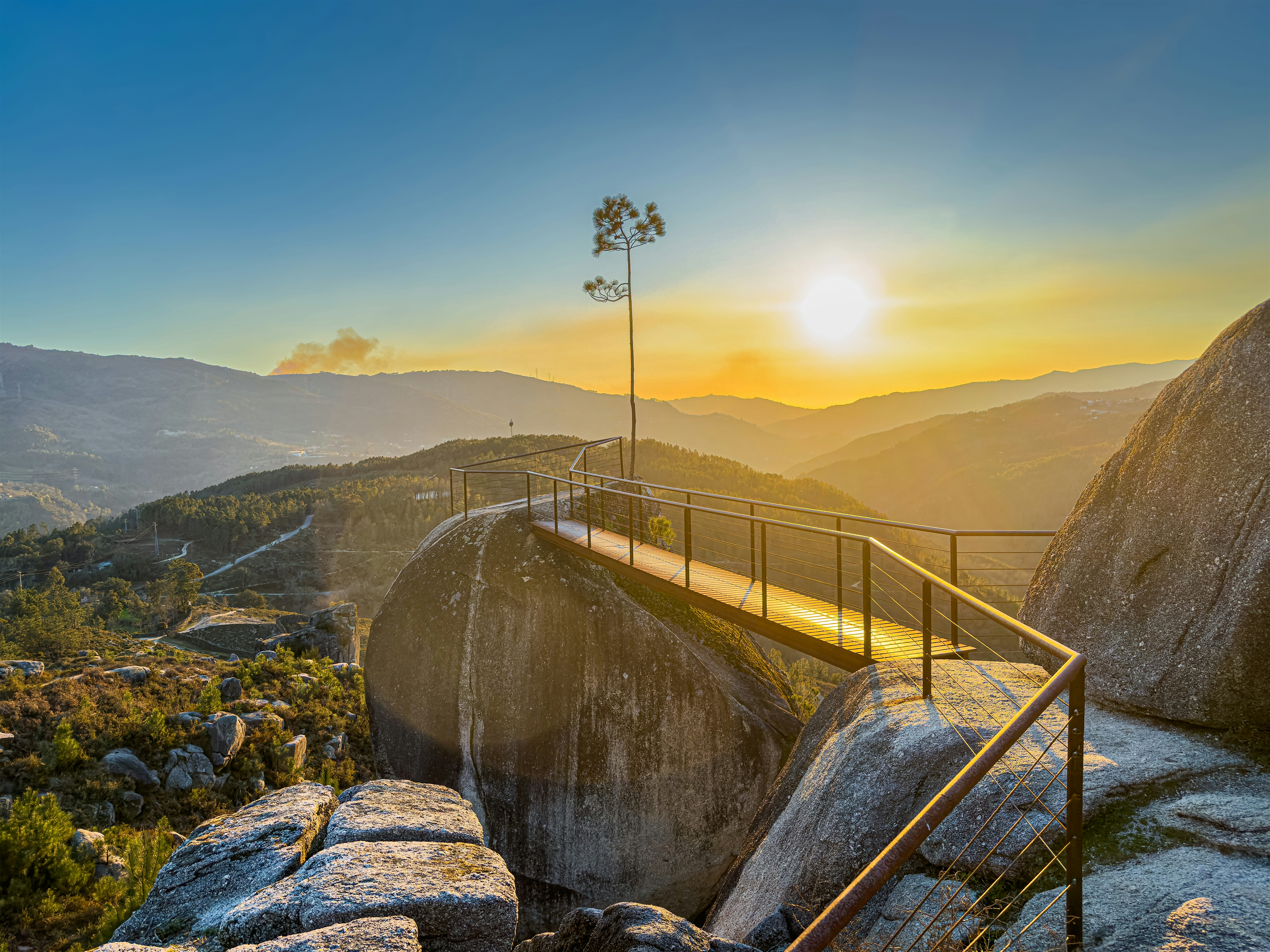 Elevated viewpoint with a walkway curving around large rocks, overlooking a mountain landscape at sunset.