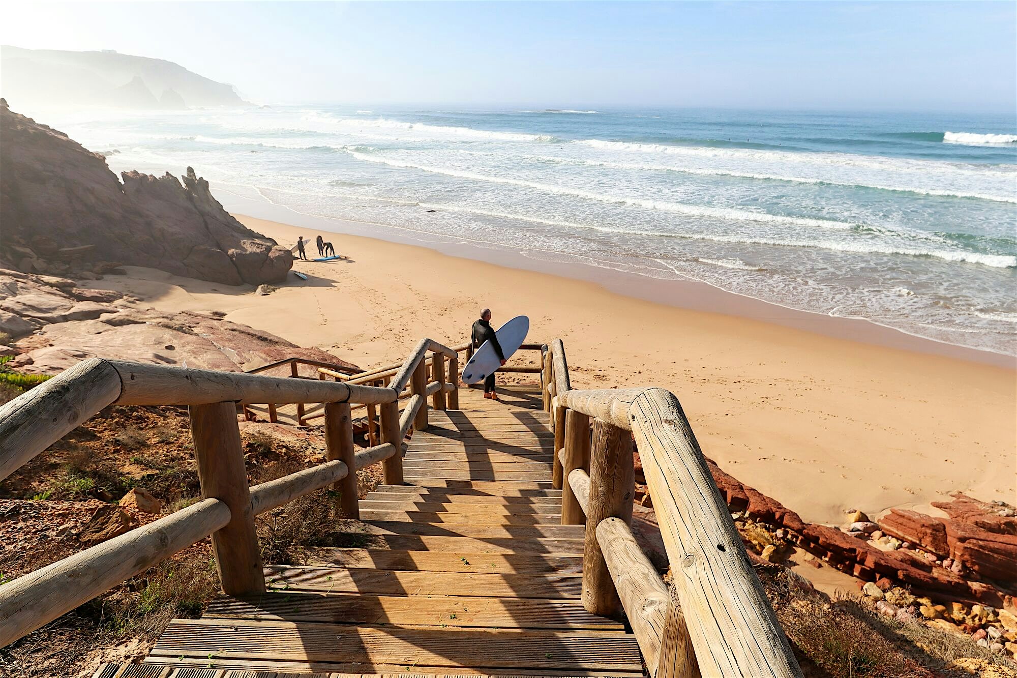 Wooden steps with railings lead down from rocky cliffs to a wide sandy beach