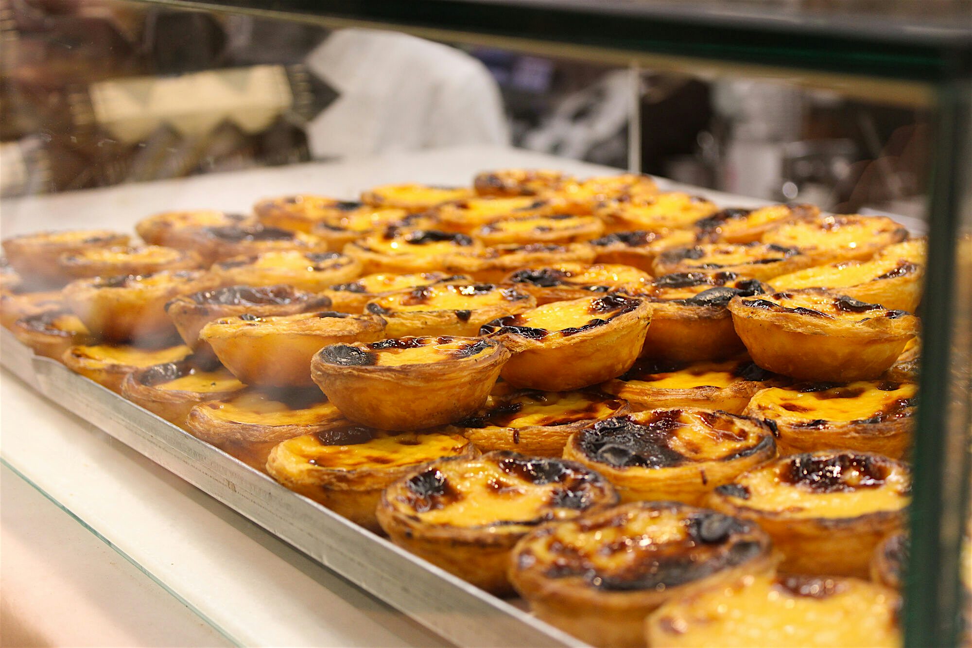 Tray of golden custard tarts with caramelised tops displayed behind a glass counter in a bakery.