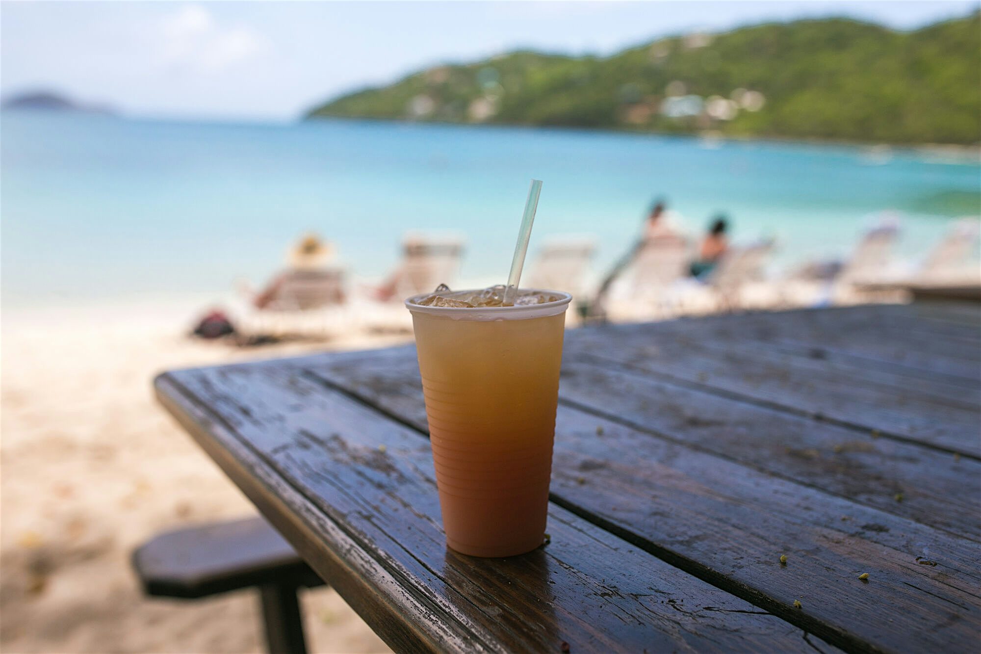 Rum Punch in a clear cup on a wooden table overlooking a sunny beach with people on sun loungers.