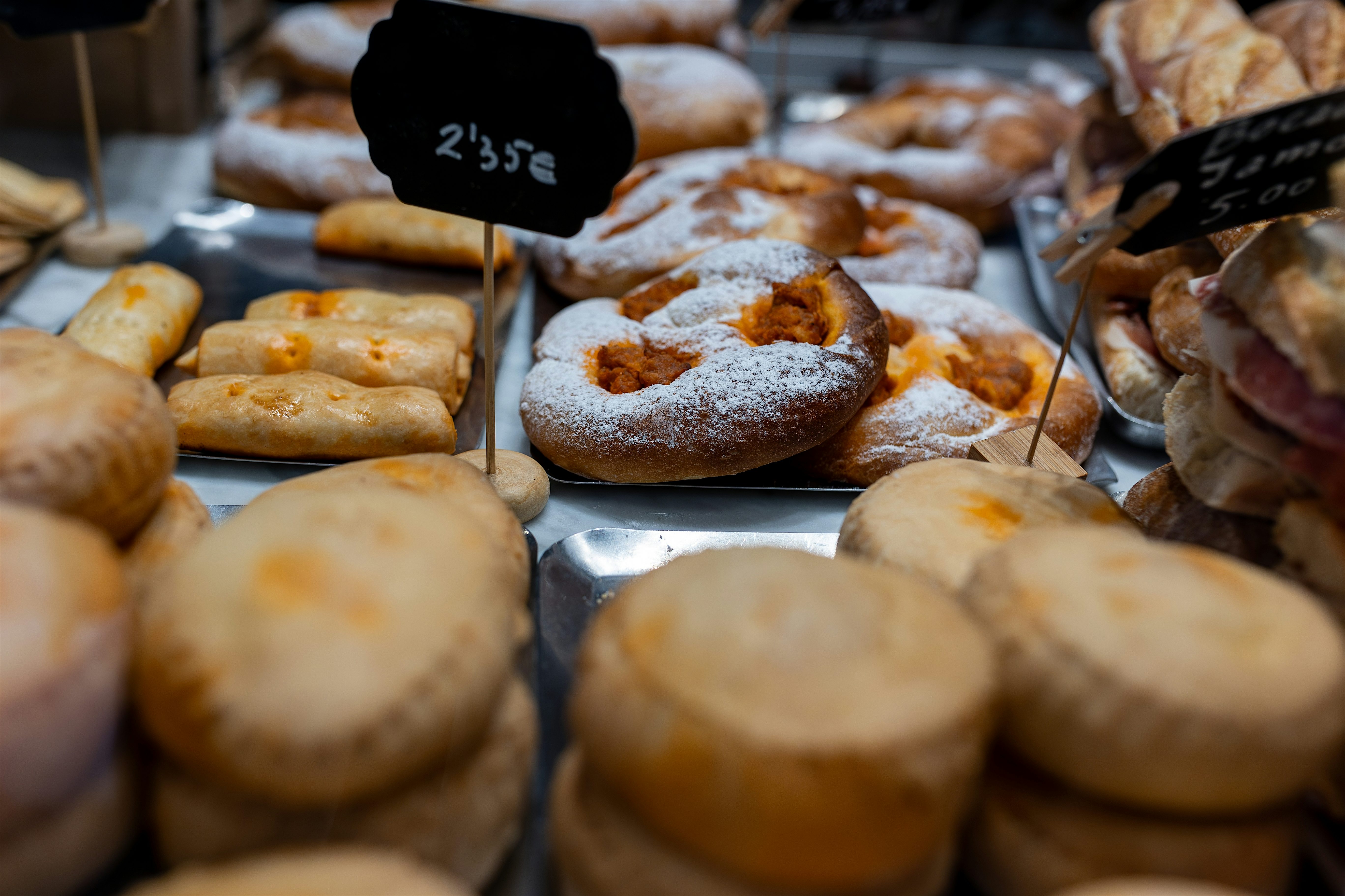 Display of traditional pastries and biscuits arranged on metal trays, dusted with sugar and labelled with small handwritten price signs.