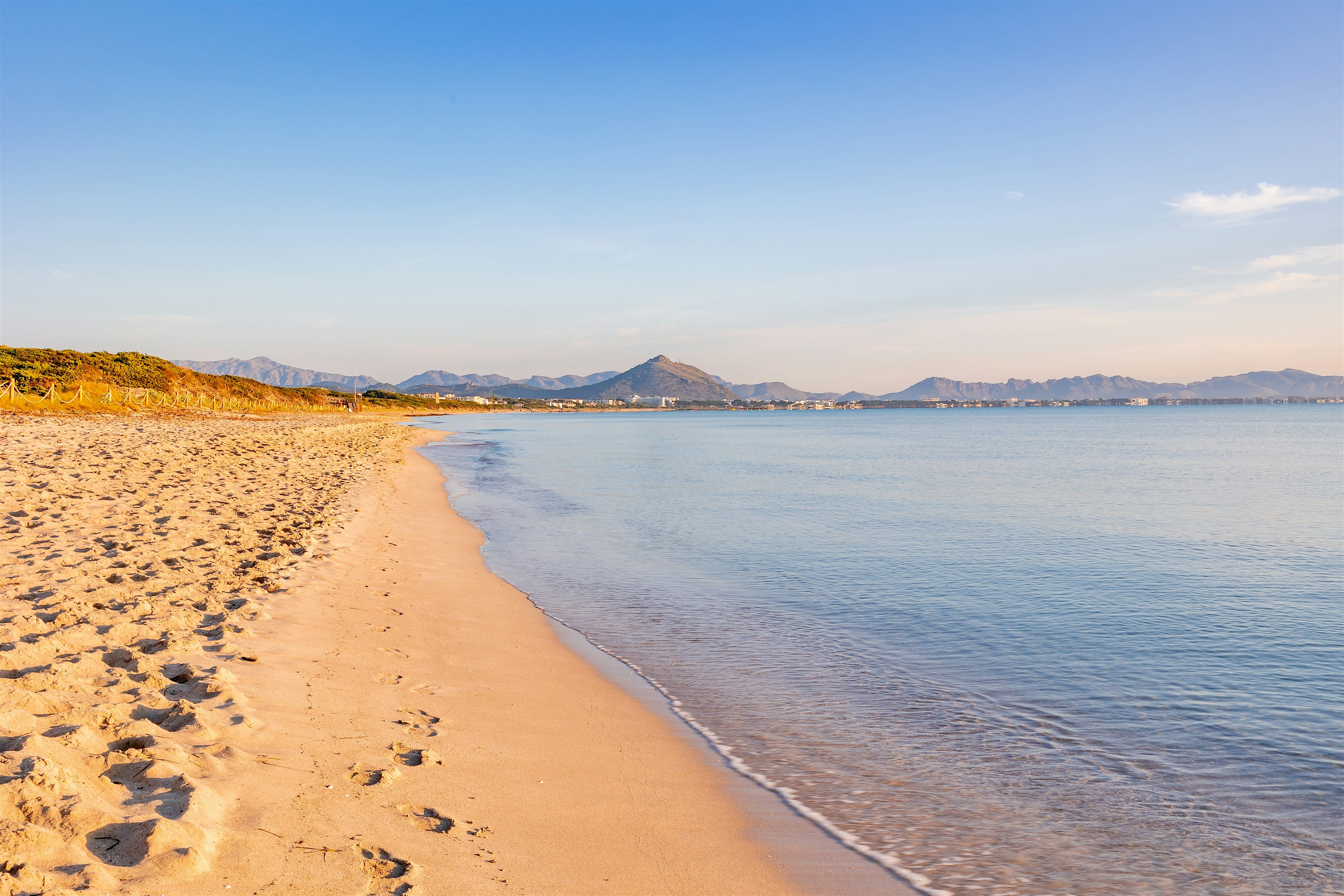 Quiet sandy beach curving along calm water, with gentle waves lapping the shore and mountains faintly visible in the distance.