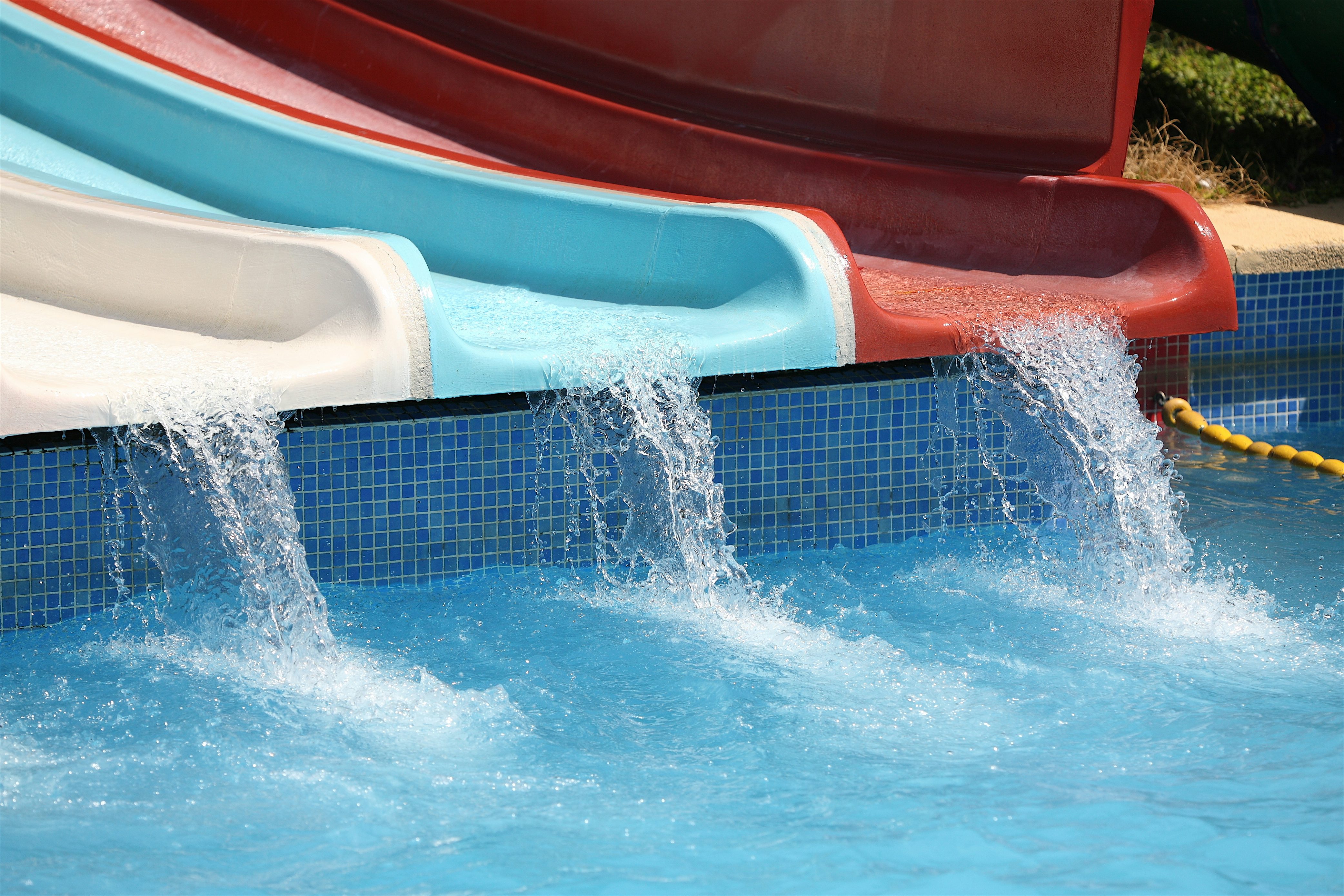 Water cascading from colourful waterslides into a bright blue pool, splashing over tiled edges in a sunny water park setting.