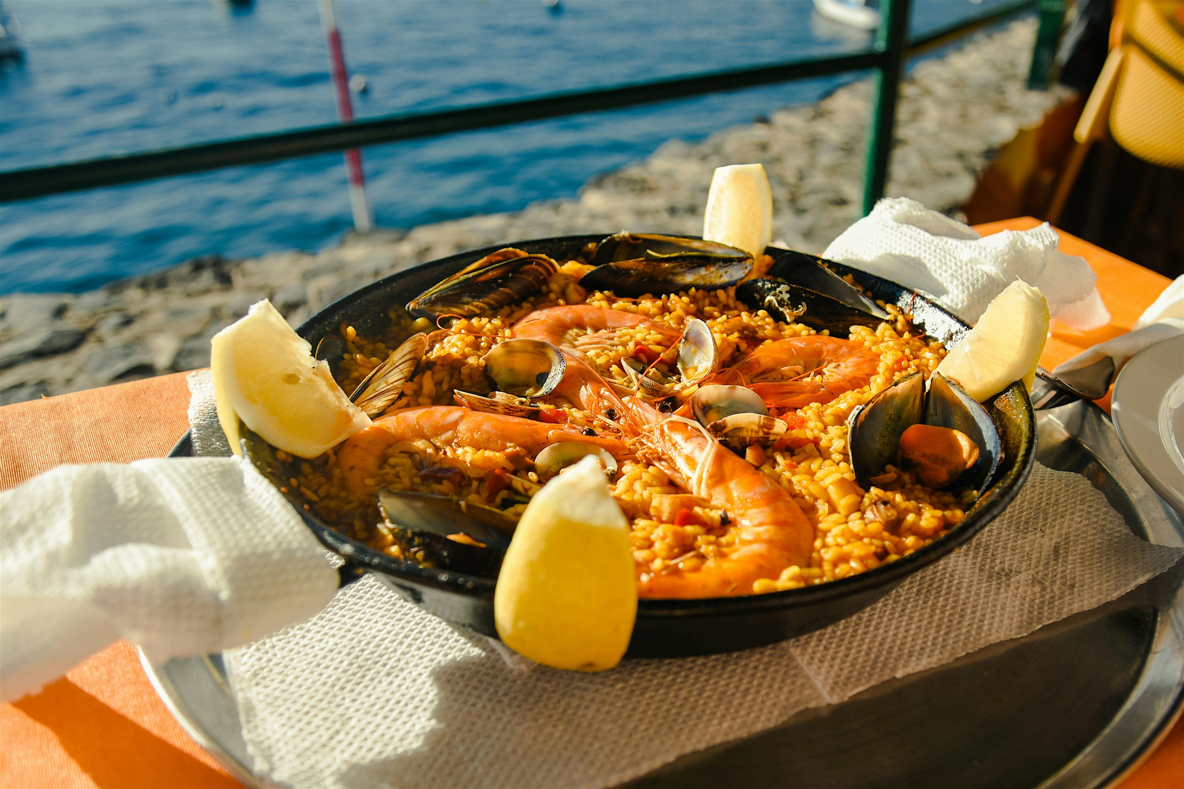 Seafood paella served in a wide black pan by the water, topped with prawns, mussels, and lemon wedges on a sunlit table.