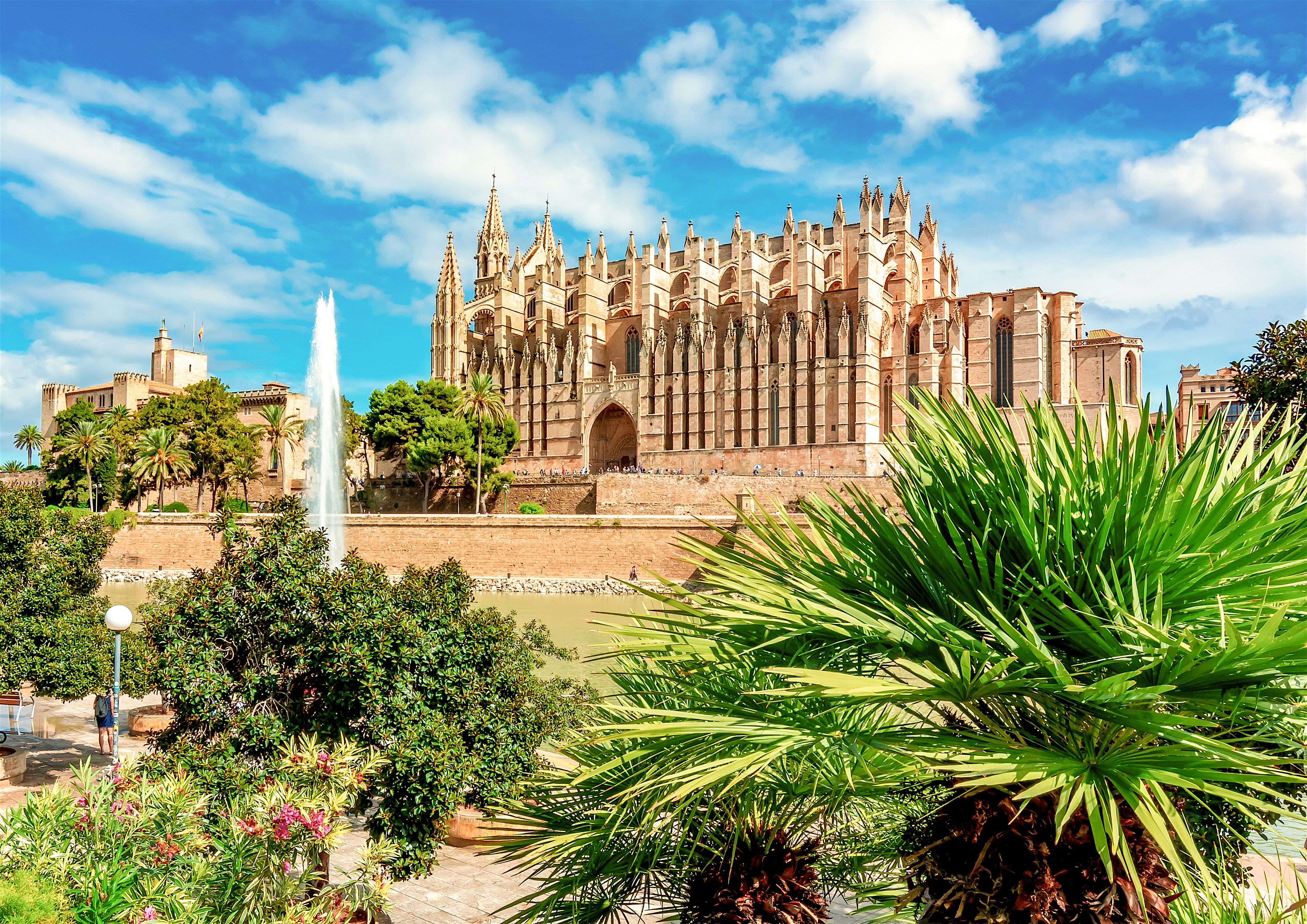 Gothic cathedral surrounded by lush gardens and palm trees, with a fountain spraying water in front under a vivid blue sky.