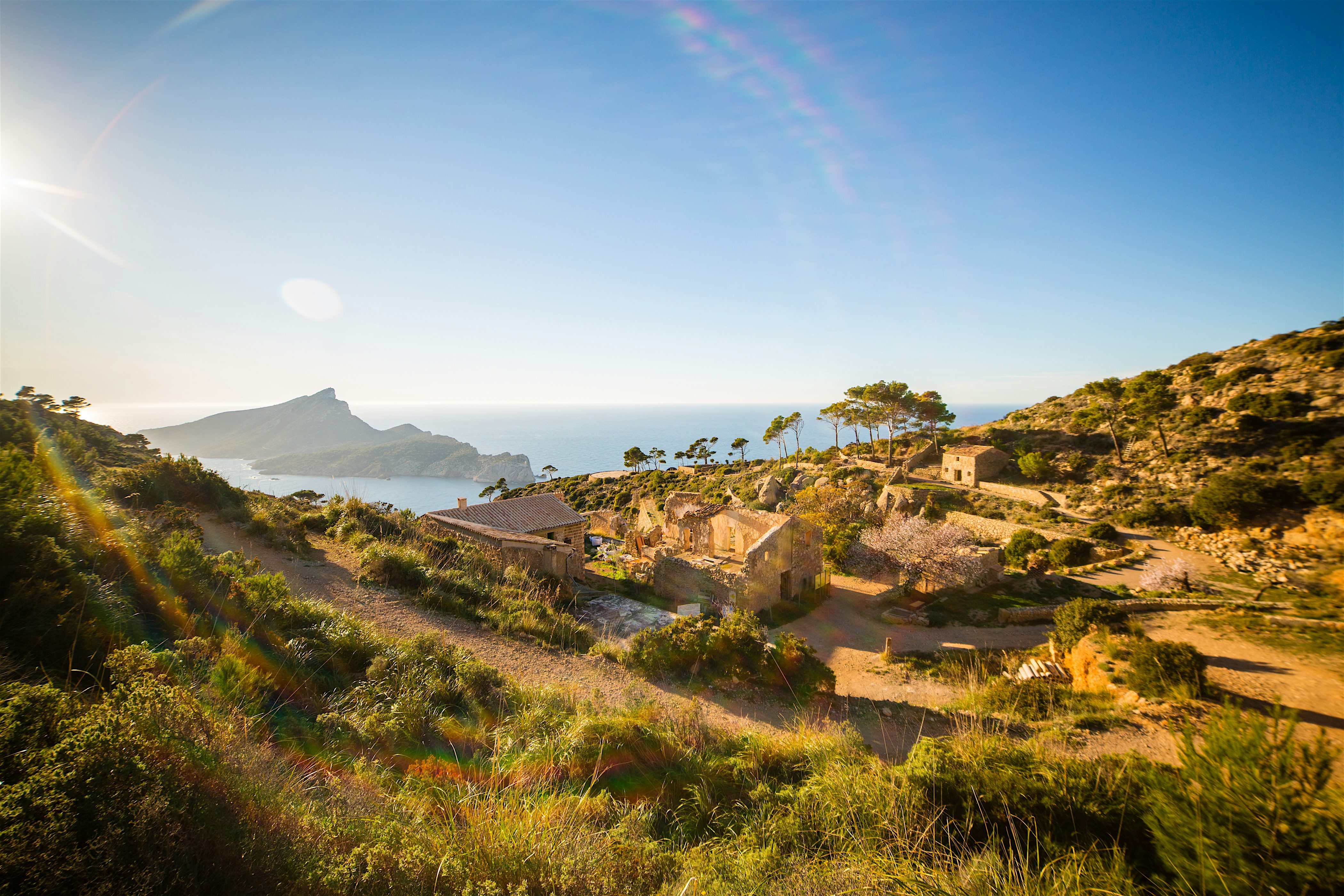 Sunlit hillside ruins overlooking the sea, surrounded by scrubland and winding paths with distant cliffs and coastline on the horizon.