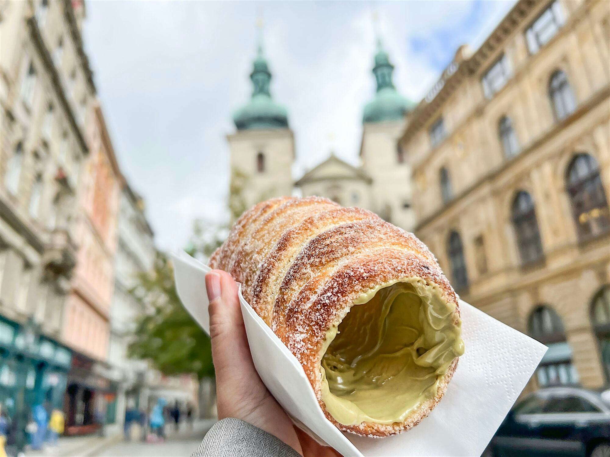 Sugar-dusted chimney cake held up on a city street, filled with thick cream and framed by historic buildings and church towers in the background.