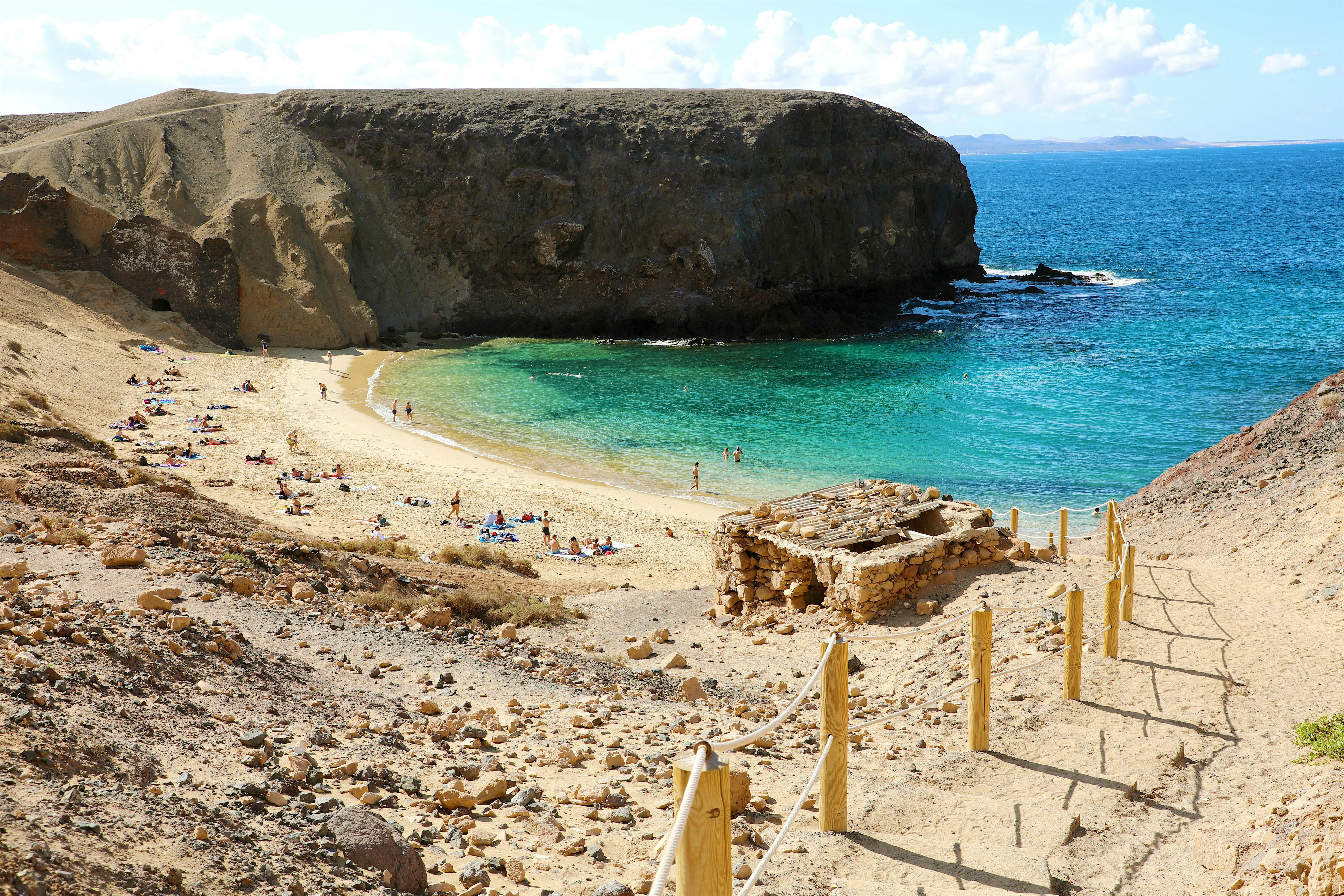 Sheltered cove with crystal-clear turquoise water and people on the small sandy beach. Large volcanic cliff to the back.