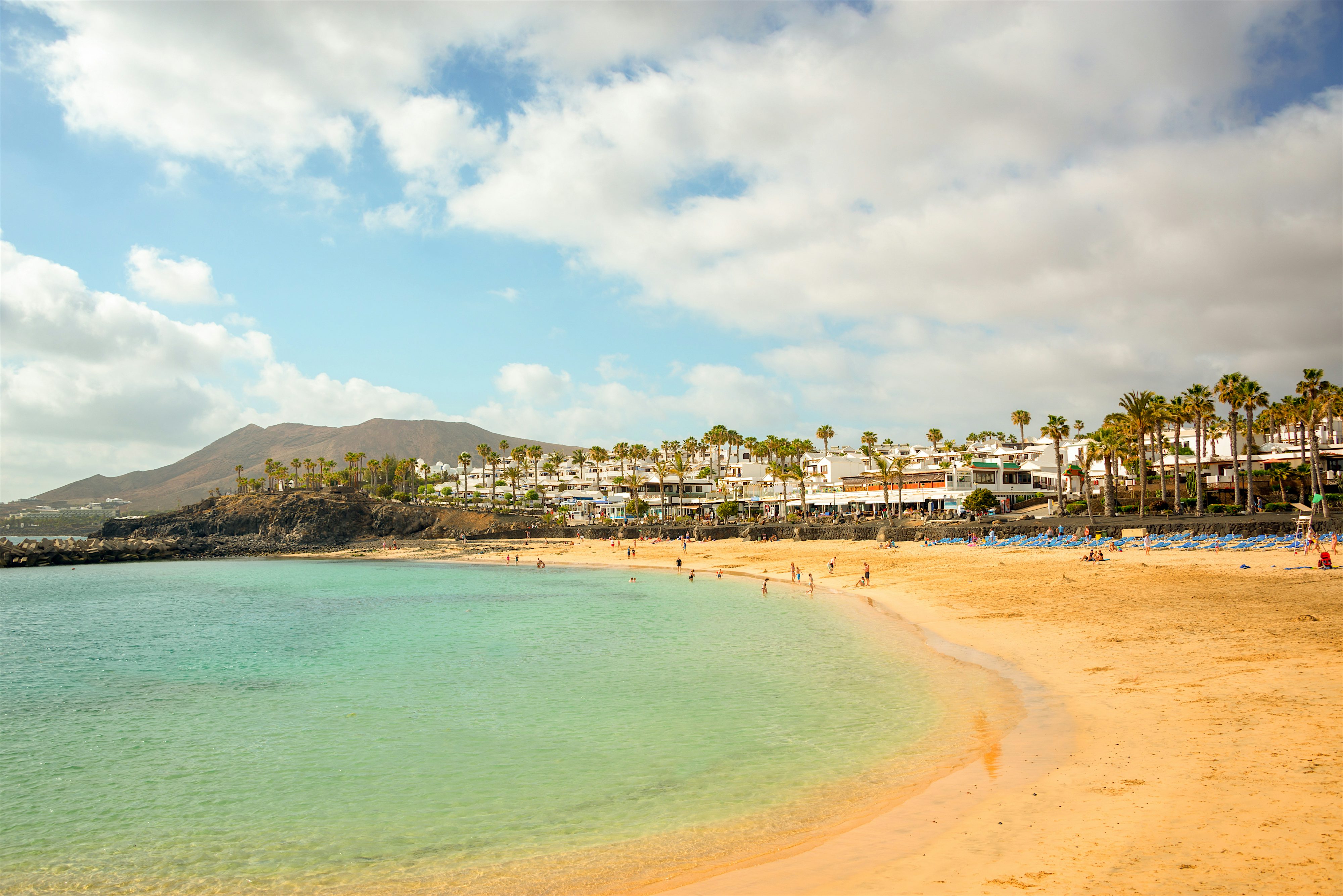 Calm turquoise water, crescent-shaped bay with restaurants, bars and hotels behind a row of palm trees. Mountain to the rear left.
