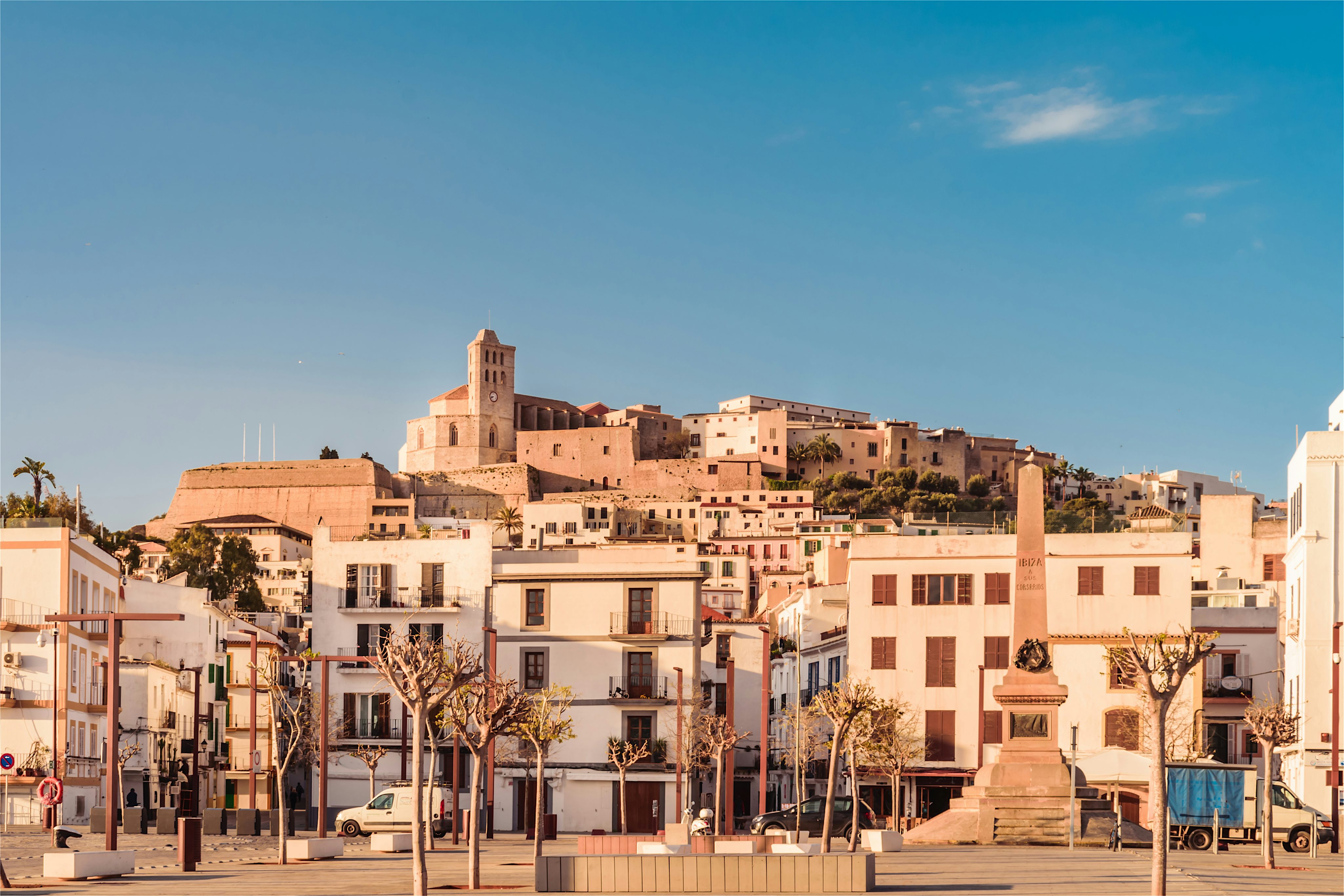 A sunlit hillside town with white buildings climbing up towards a historic church, overlooking a wide open square lined with bare trees.