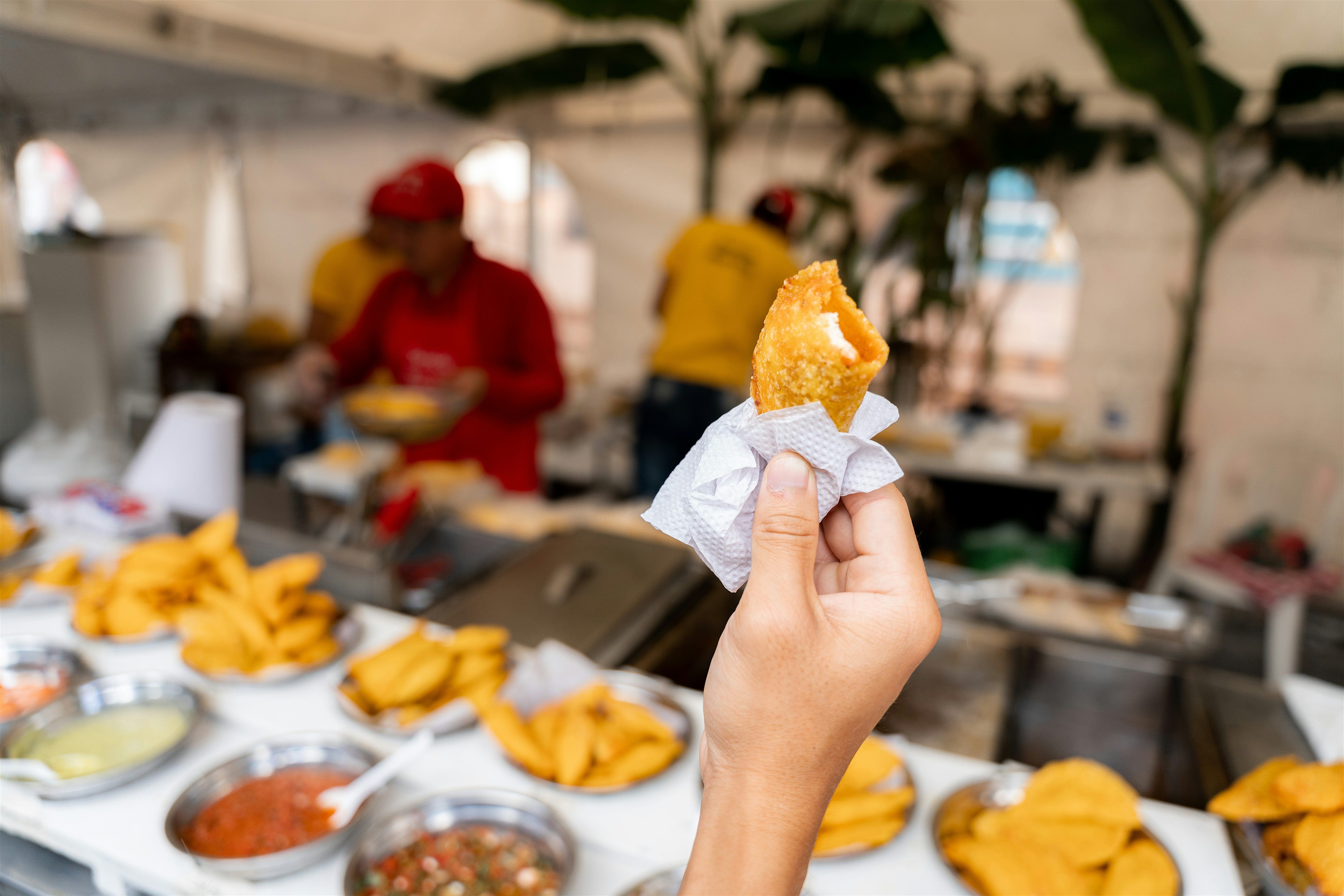Hand holding a fried snack over a street food counter, with trays of foods and bowls of dips, while vendors prepare food in the background.