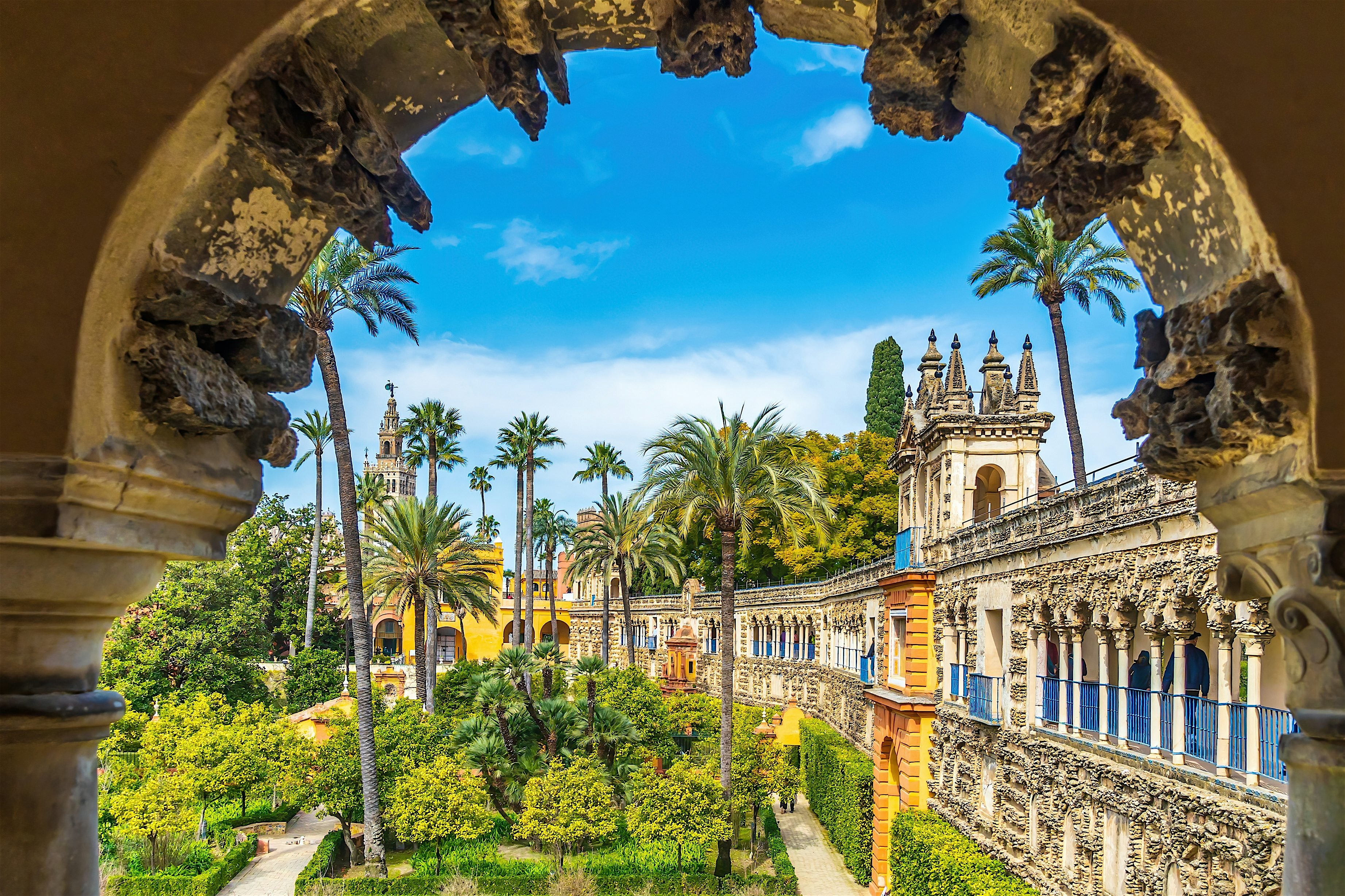 View through a stone arch into a courtyard with palm trees, formal gardens, and decorated historic buildings under a bright blue sky.