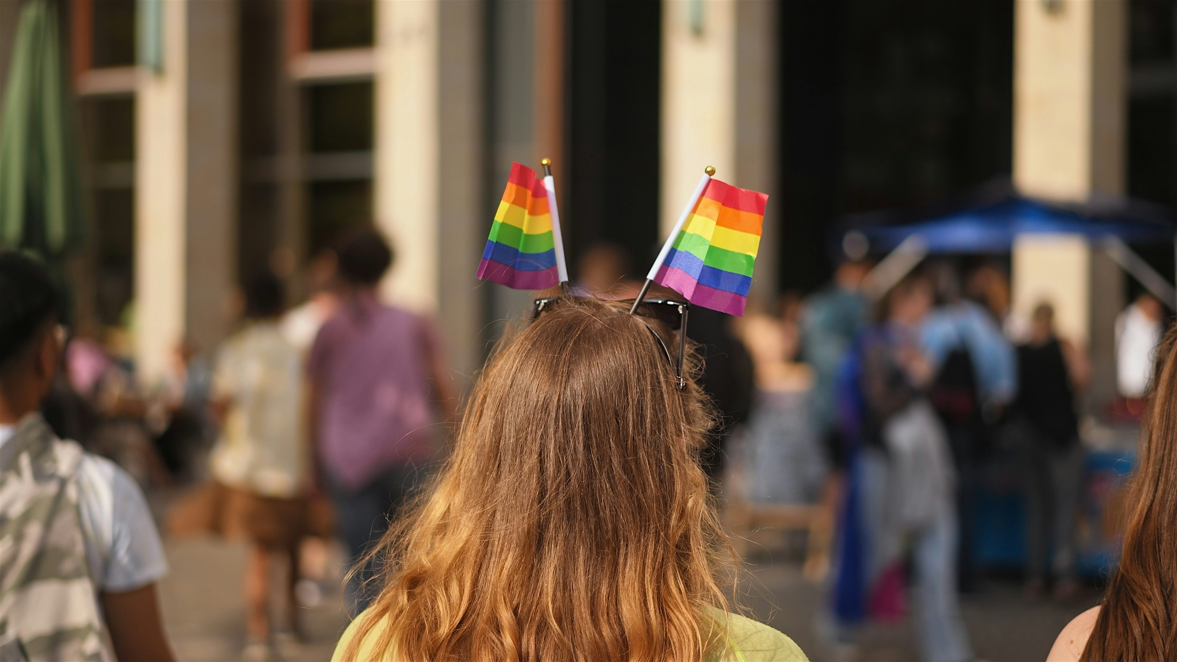 A person seen from behind wearing small rainbow flags on a headband, walking through a busy outdoor area with stalls and people in the background.