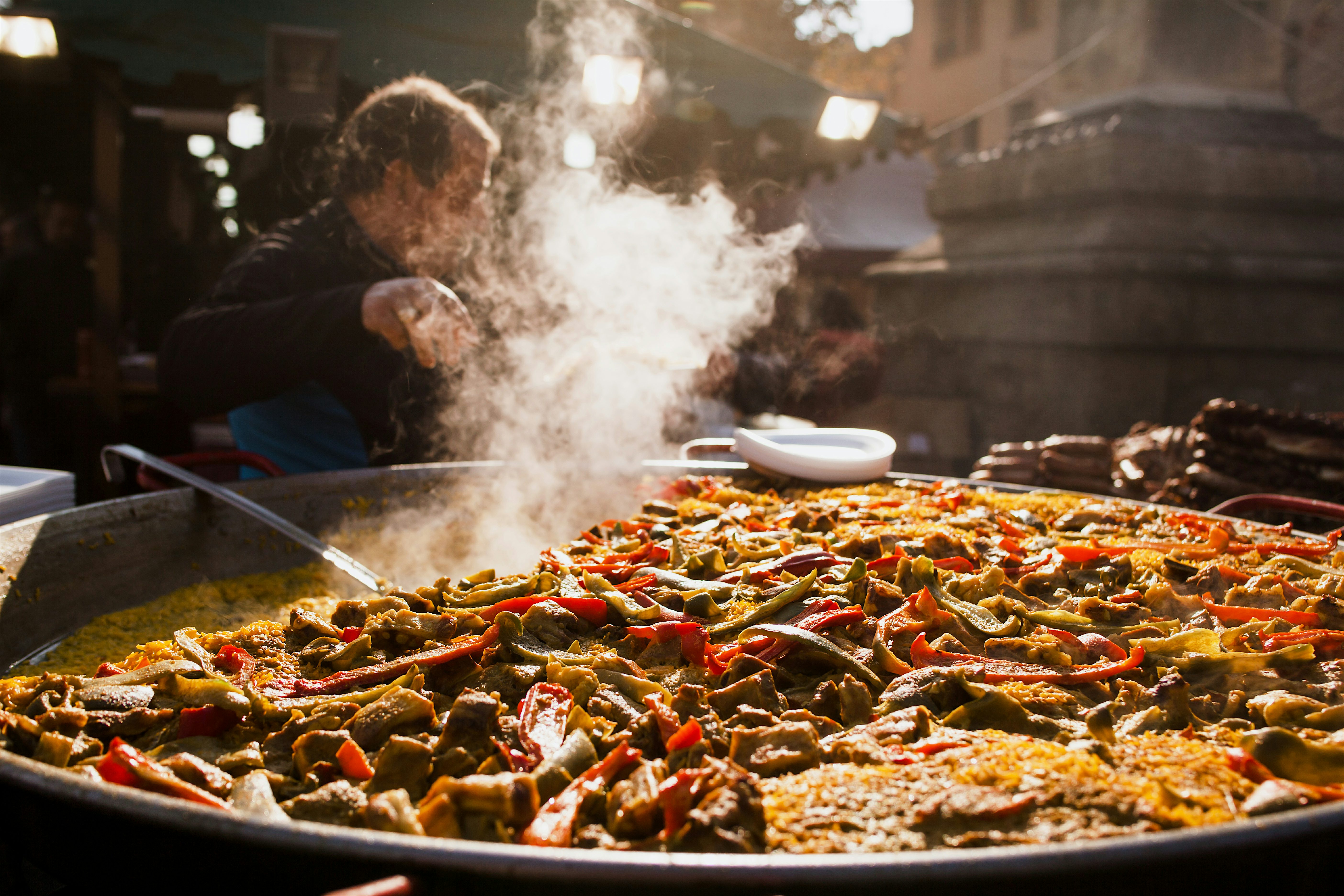 A large paella pan cooking at an outdoor market, filled with rice, vegetables, and meat, with steam rising as it’s stirred in a busy street setting.