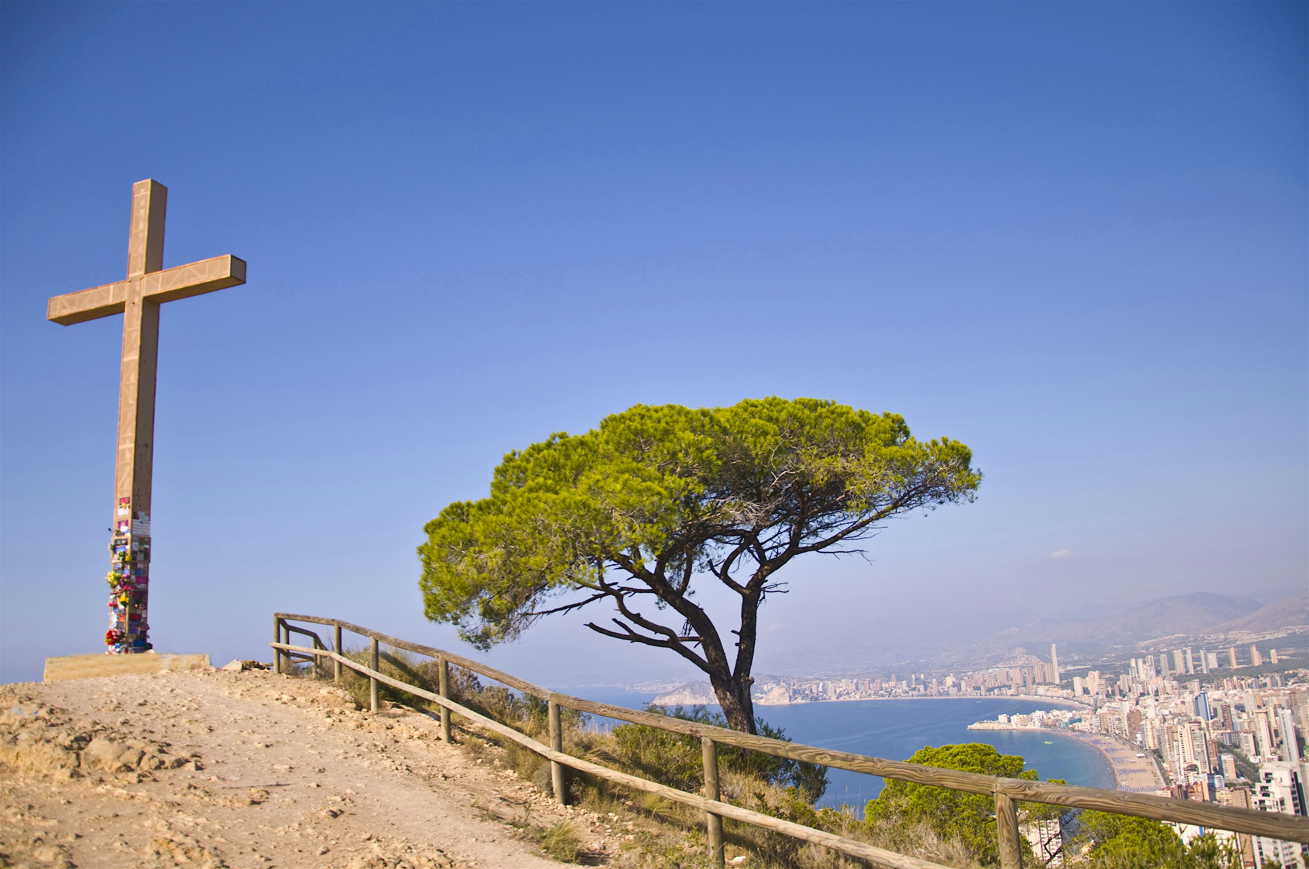 A hilltop viewpoint with a wooden cross and lone tree overlooking a coastal city and sweeping bay in the distance.