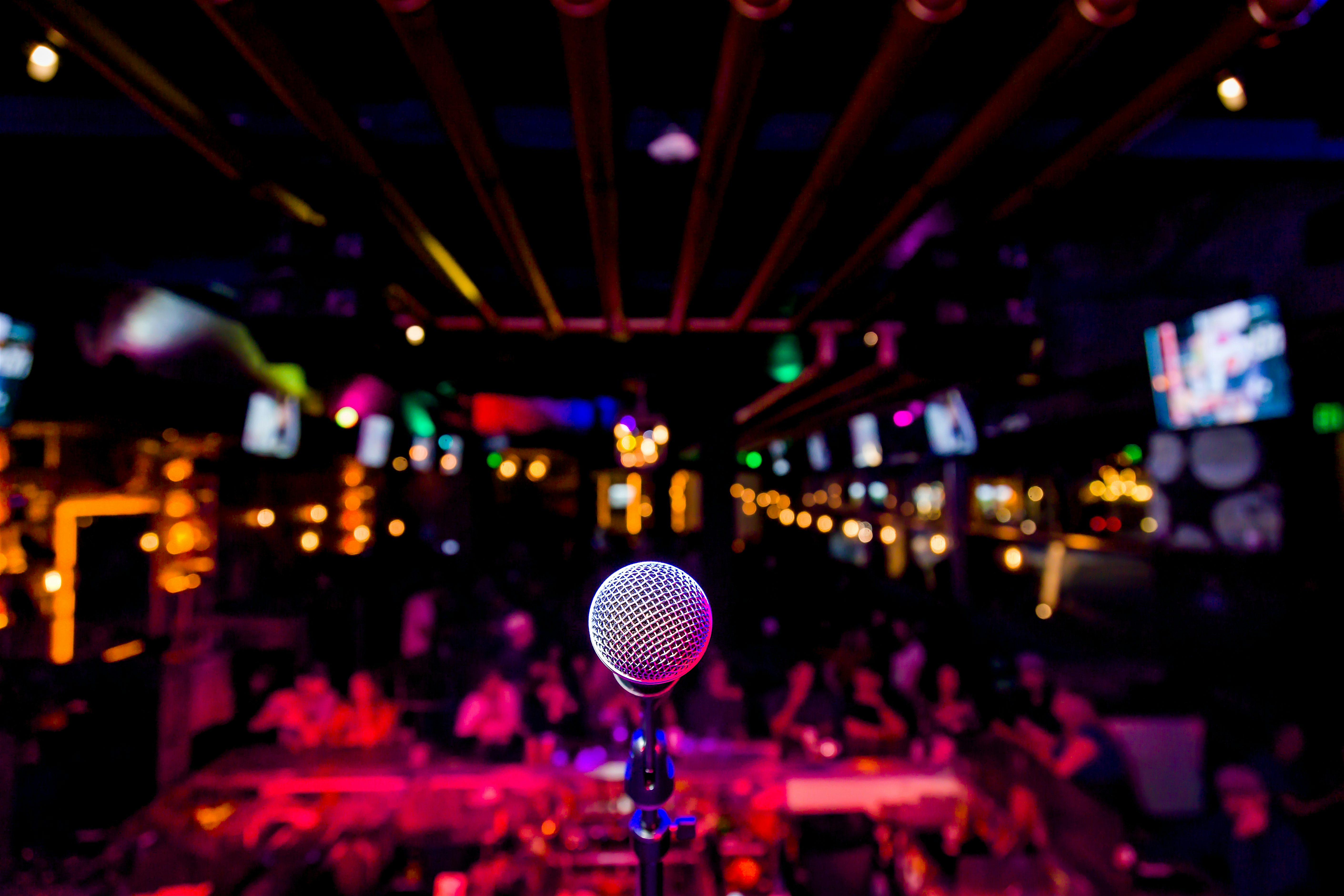 A microphone positioned on a stage facing a seated audience, with colourful club lighting and TV screens illuminating a lively indoor venue.