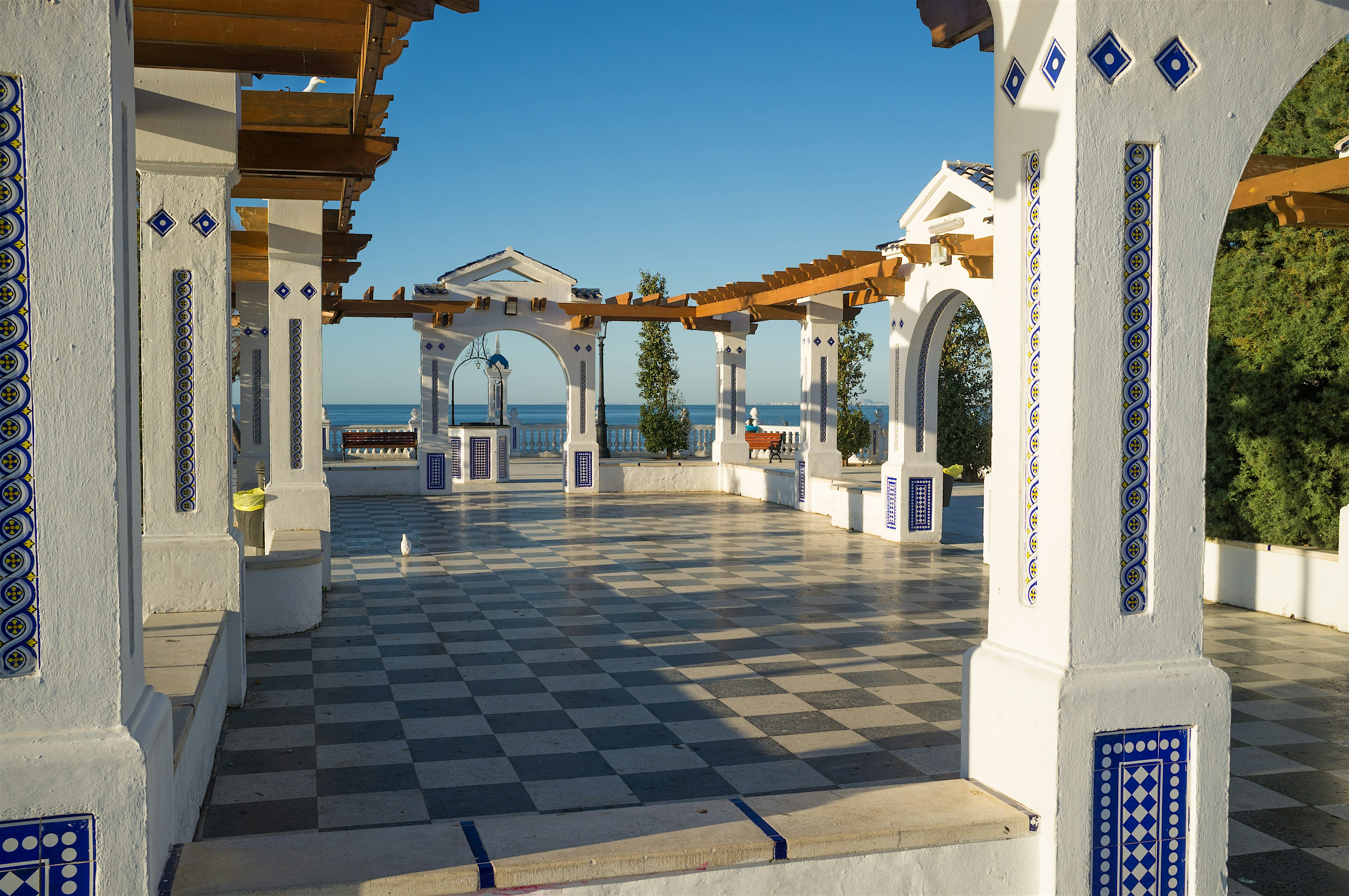 A tiled seaside terrace with white arches and decorative blue details, opening out towards the sea under a clear blue sky.