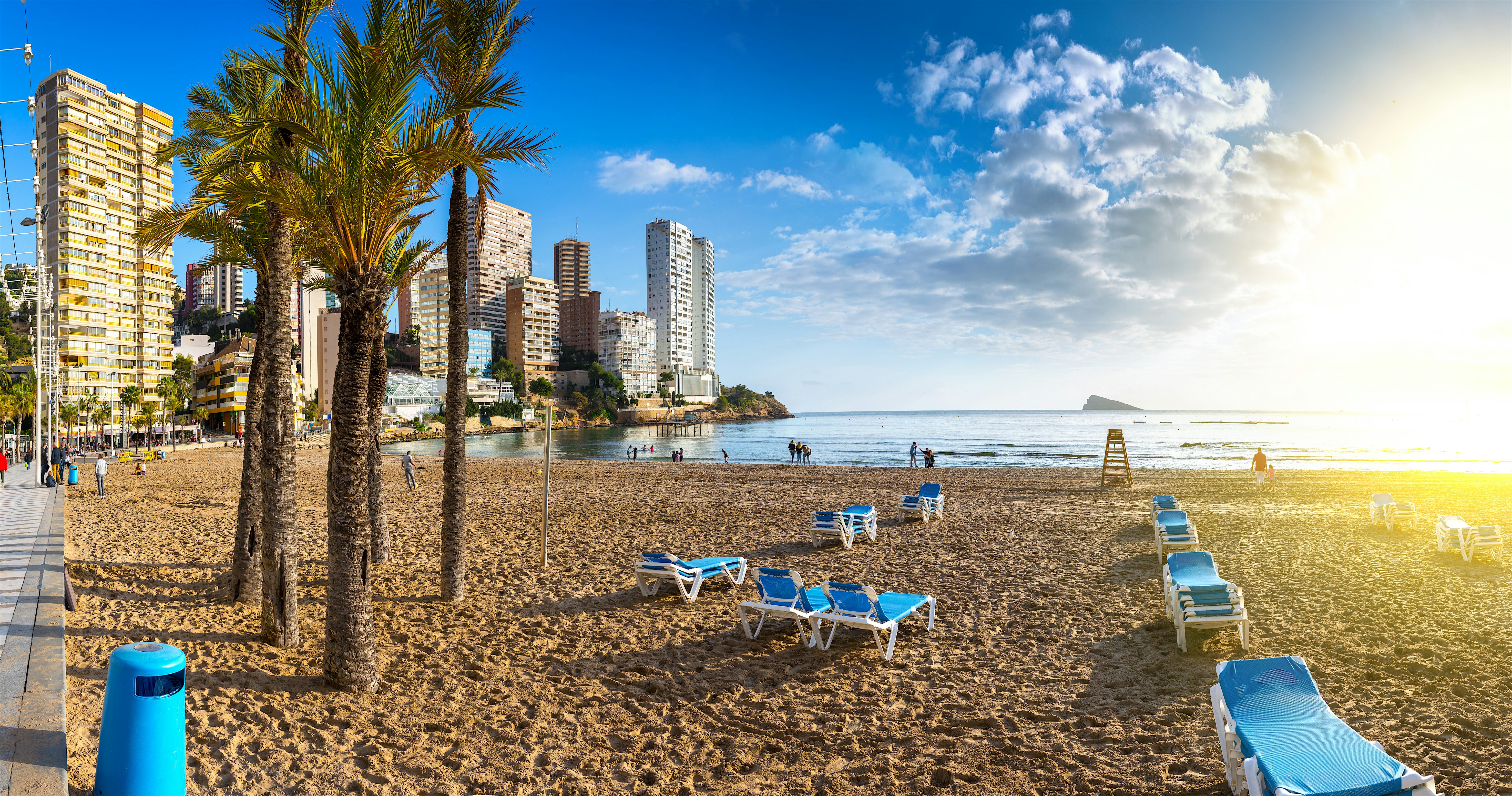 Palm-lined sandy beach with sun loungers facing calm sea water, backed by high-rise buildings along the shoreline in warm evening light.