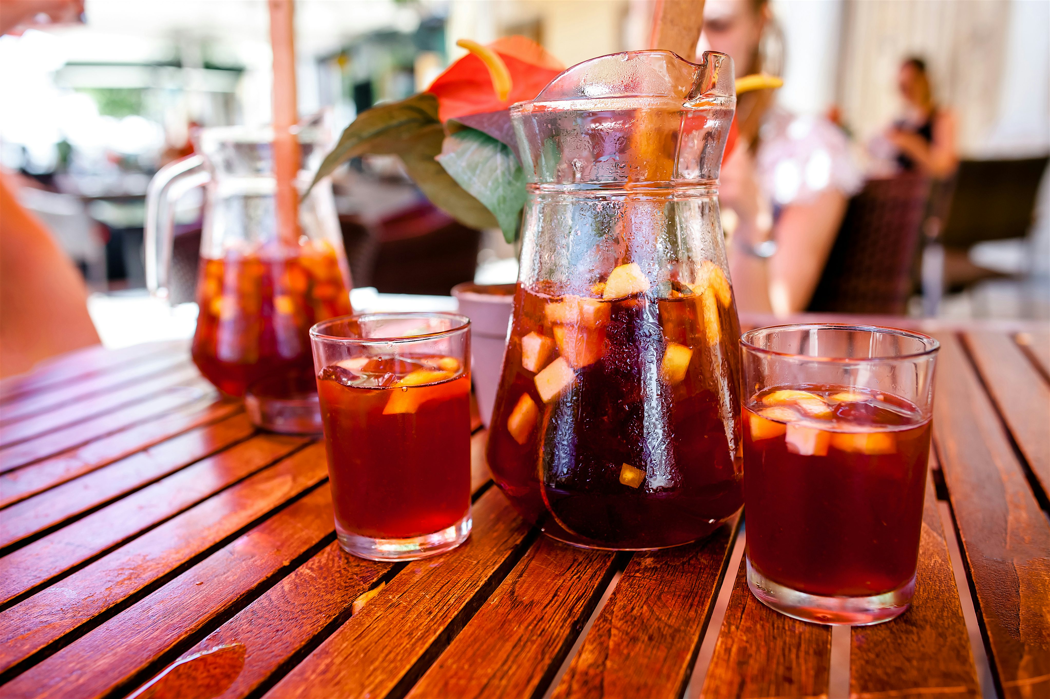 Glass jugs and tumblers of sangria on a wooden table, filled with fruit pieces and ice in a relaxed café environment.