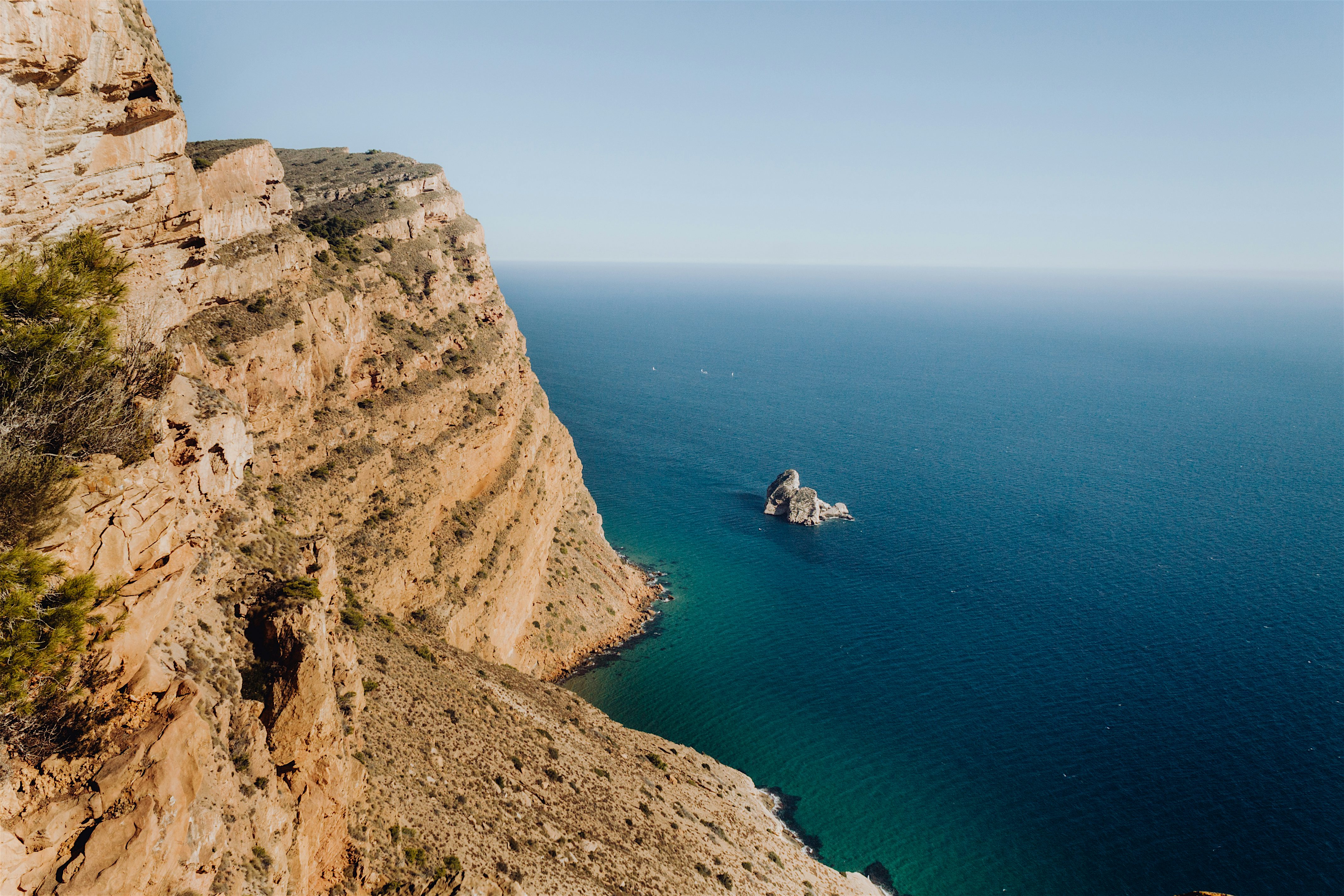 A rugged coastal cliff dropping into deep blue sea, with layered rock formations and a small rocky outcrop offshore under a clear sky.