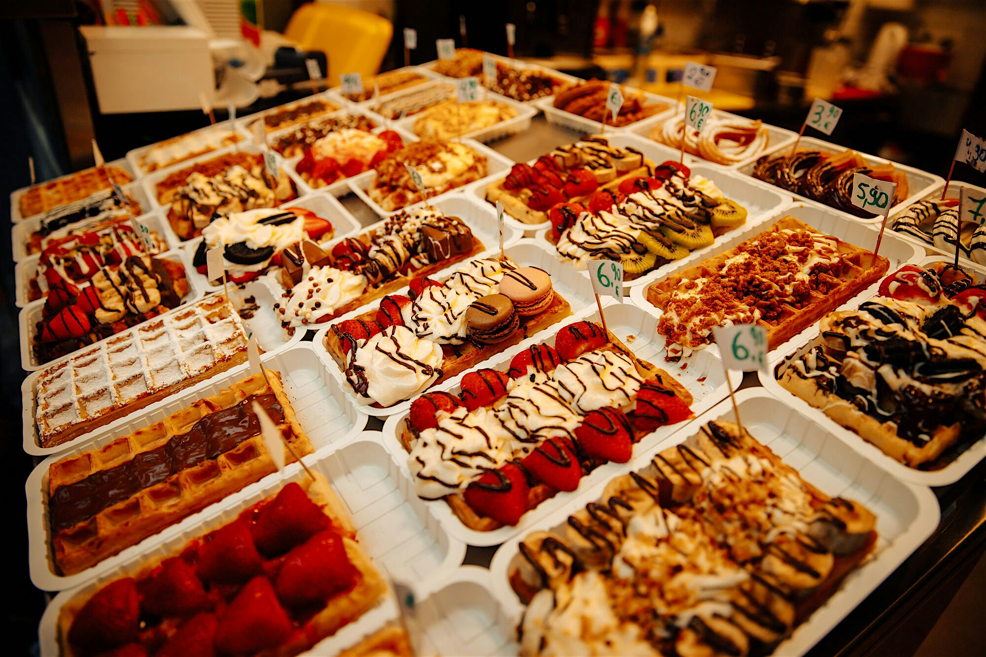 Selection of freshly made waffles topped with strawberries, chocolate drizzle, whipped cream, and assorted toppings, neatly arranged behind a glass counter with small price tags.