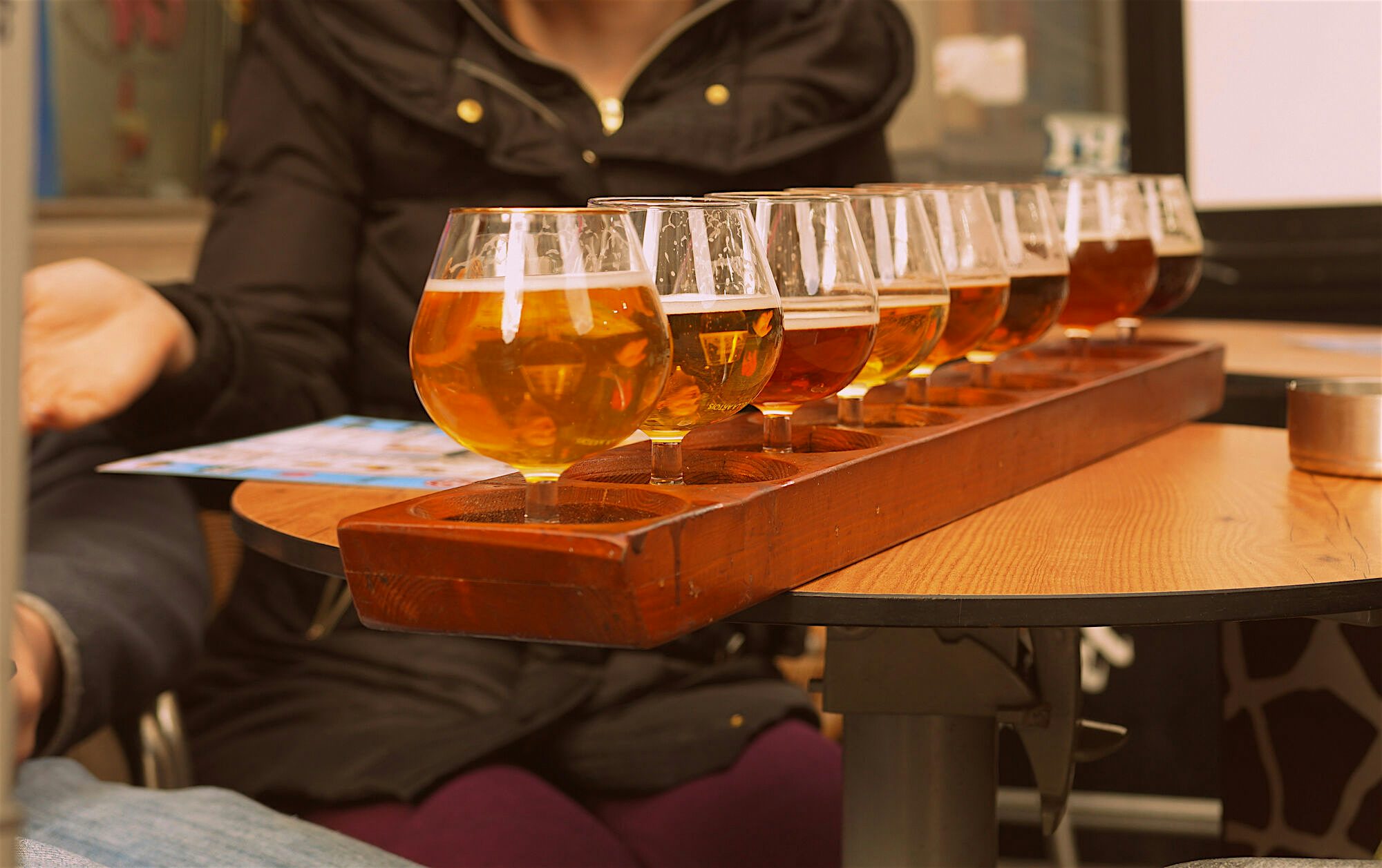 Row of small beer glasses arranged on a wooden tasting board, served at a table for a beer flight.