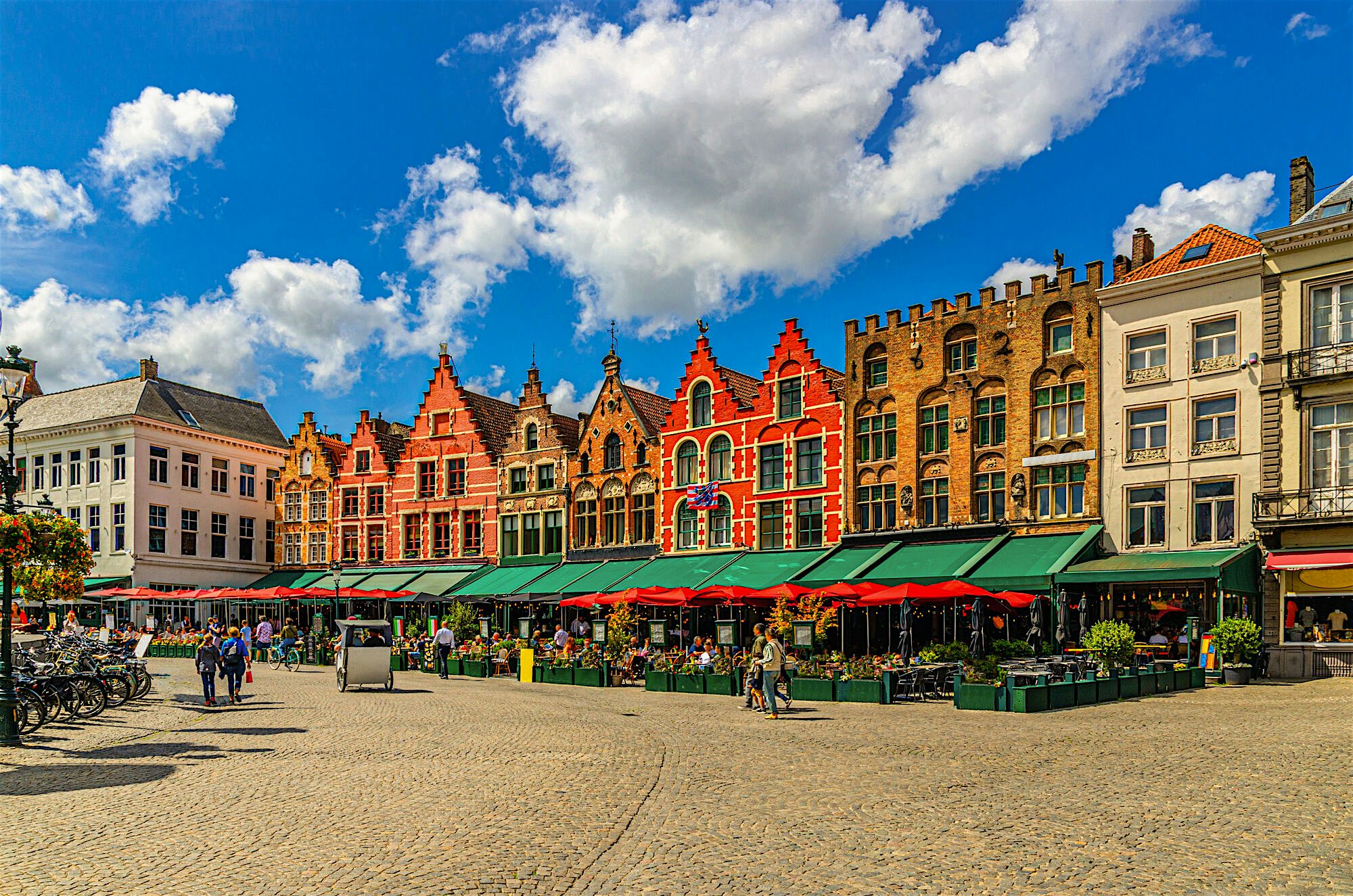 Lively market square surrounded by stepped-gable buildings, outdoor cafés spilling into an open cobbled plaza under a bright blue sky.