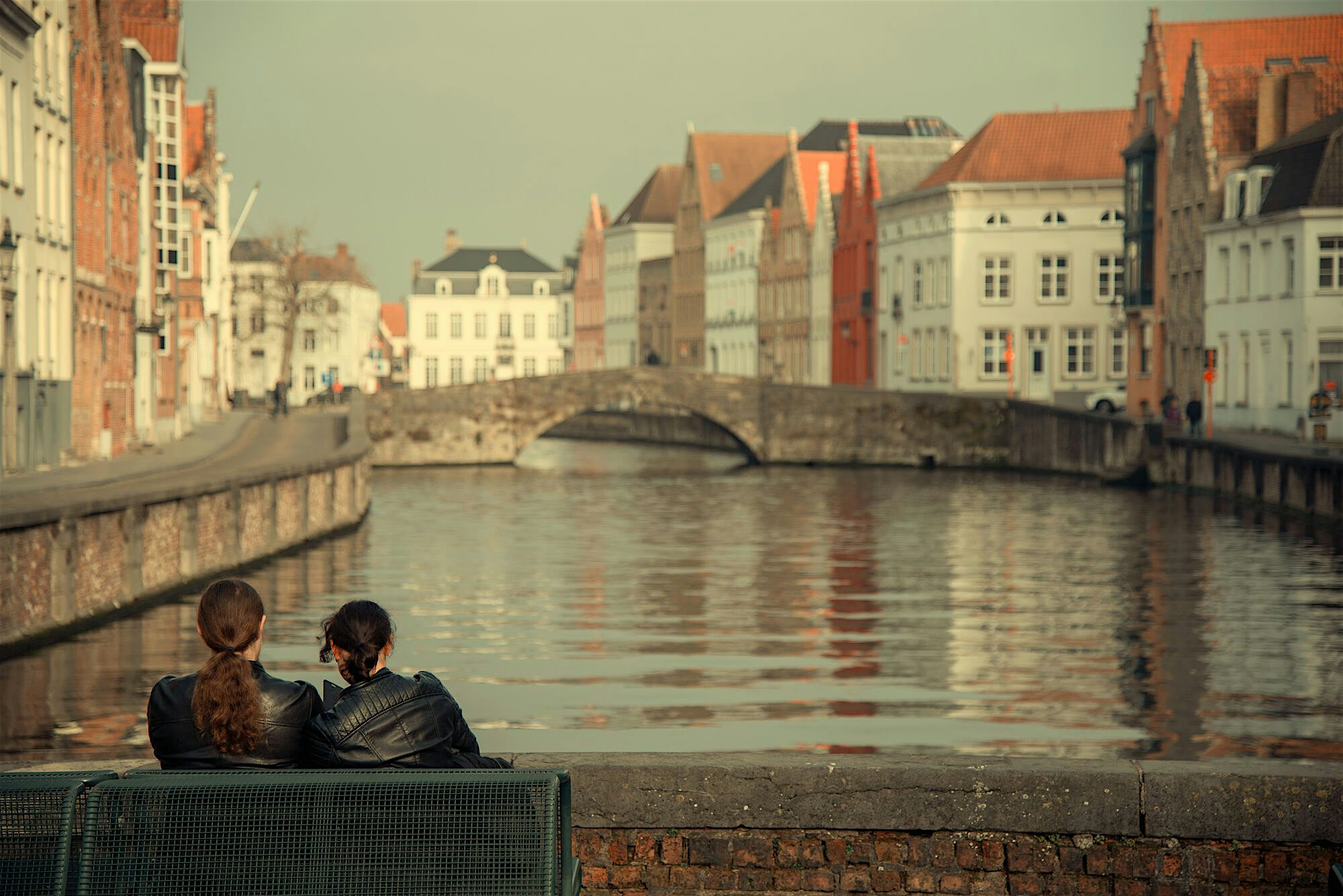 Two people sitting on a bench beside a calm canal, looking out over water lined with historic buildings and a stone bridge.