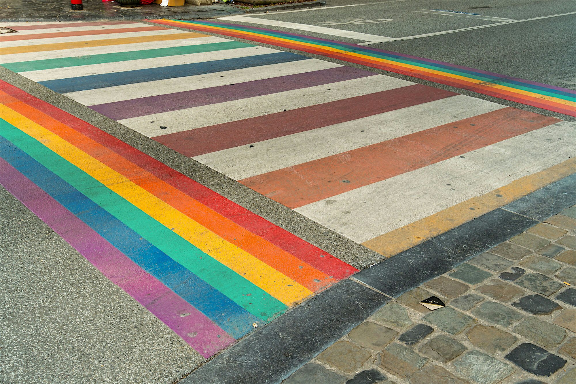 Rainbow-painted pedestrian crossing stretching across a city street, bordered by stone paving and road markings.