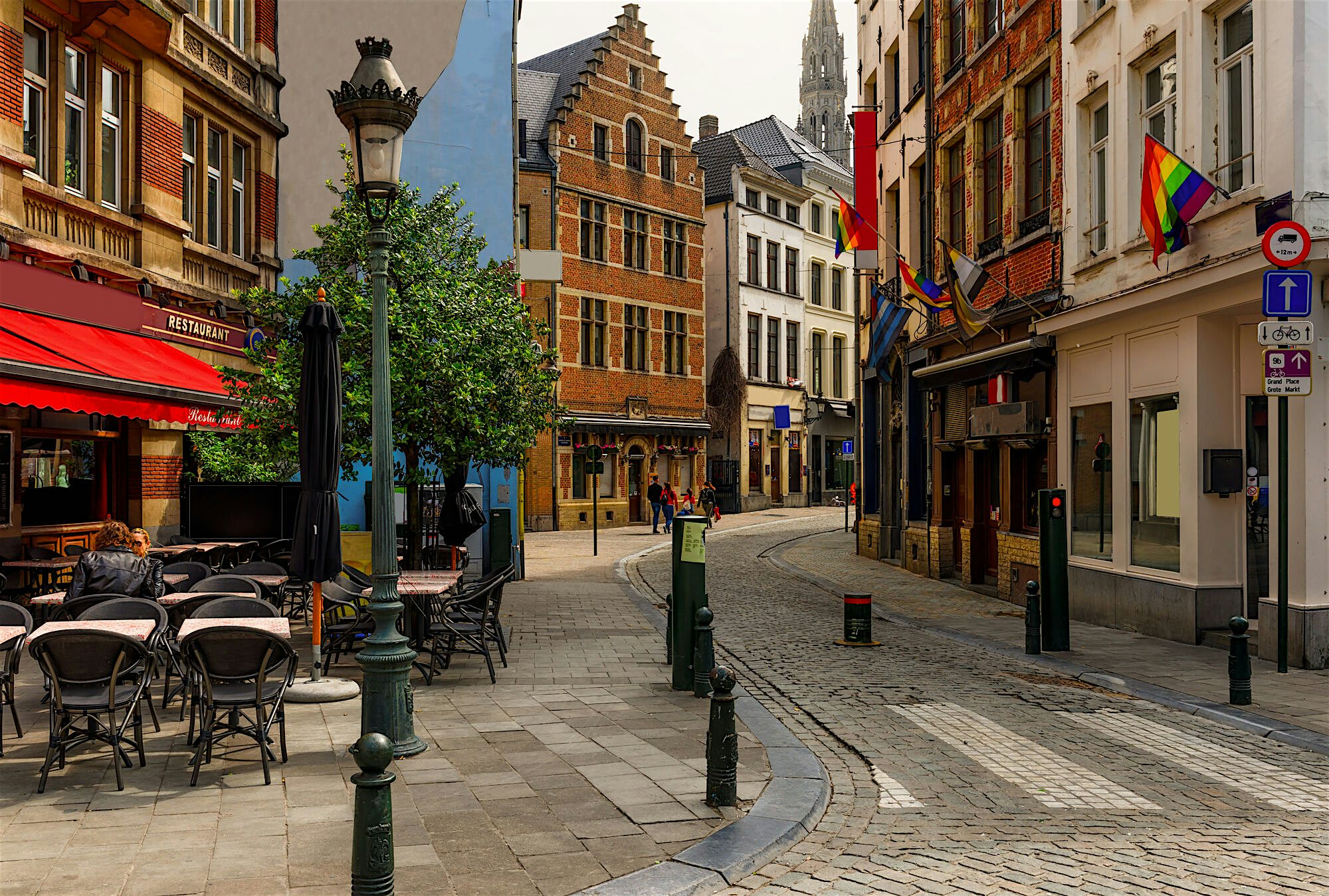 Narrow cobbled street with outdoor café seating, historic brick buildings, and rainbow flags hanging from façades.