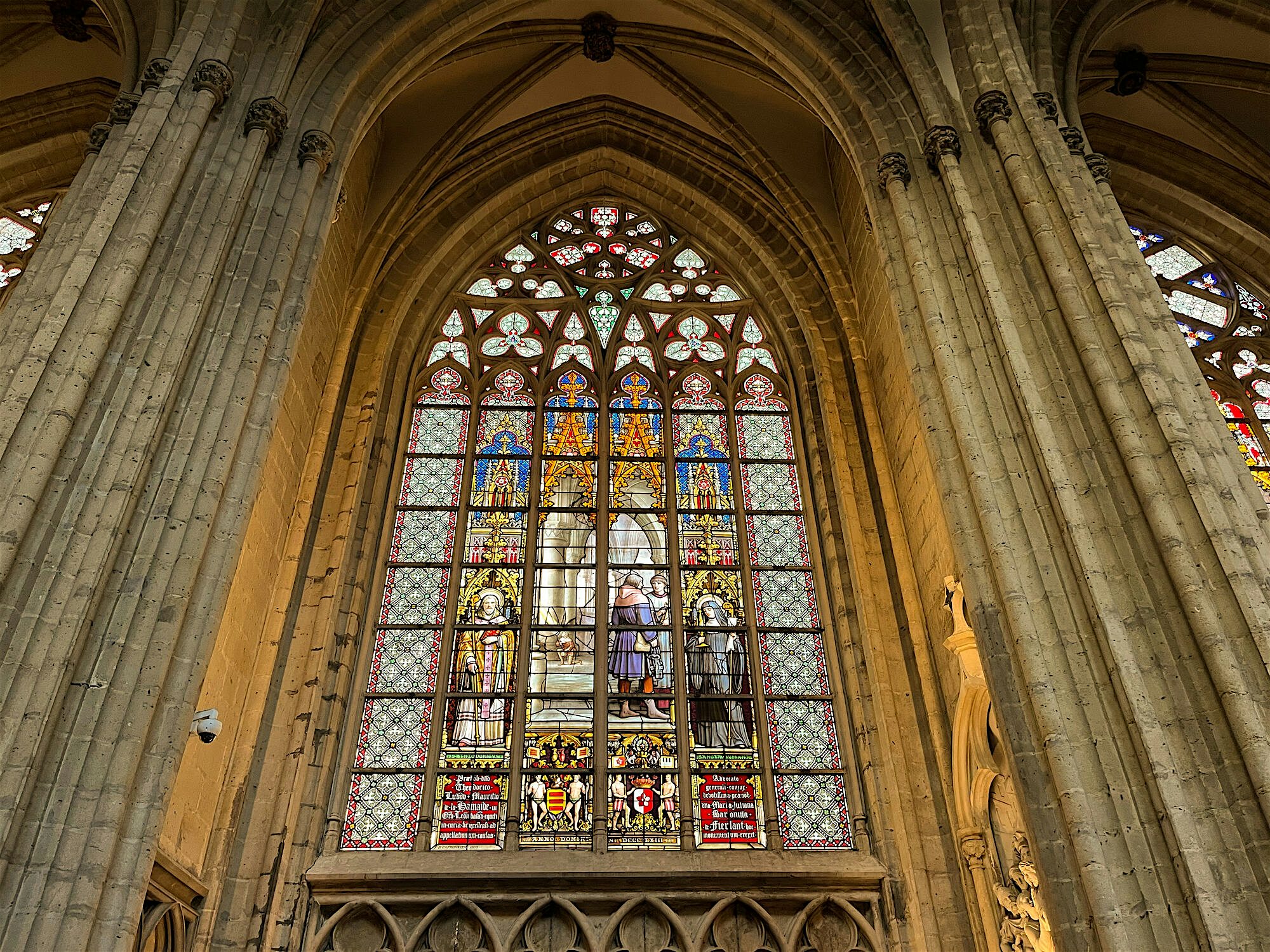 Large stained-glass window set within towering stone arches, filled with colourful religious figures and intricate patterns inside a historic church.