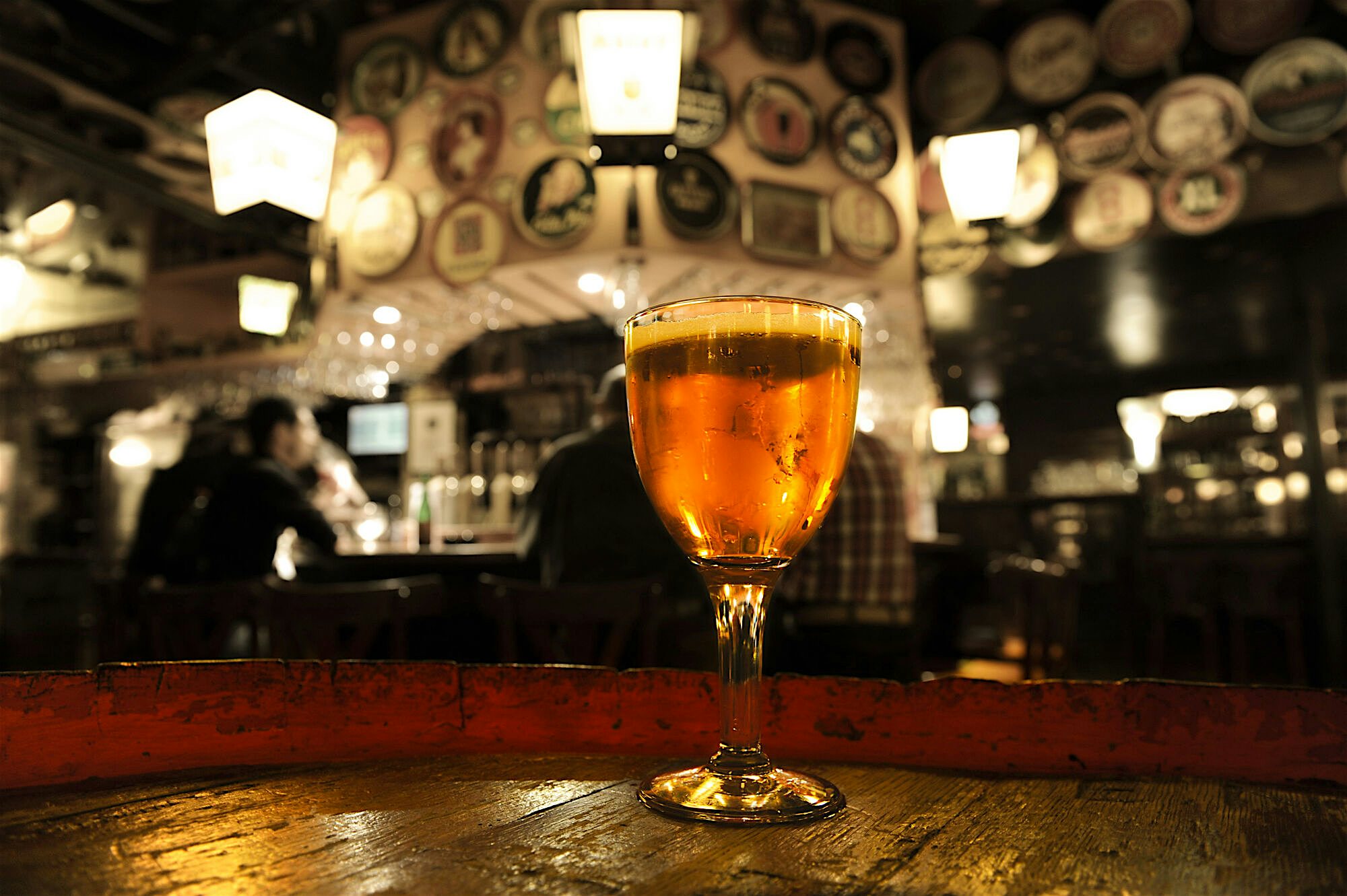 Glass of amber beer resting on a rustic wooden table inside a cosy bar, with warm lighting and walls covered in decorative plates in the background.
