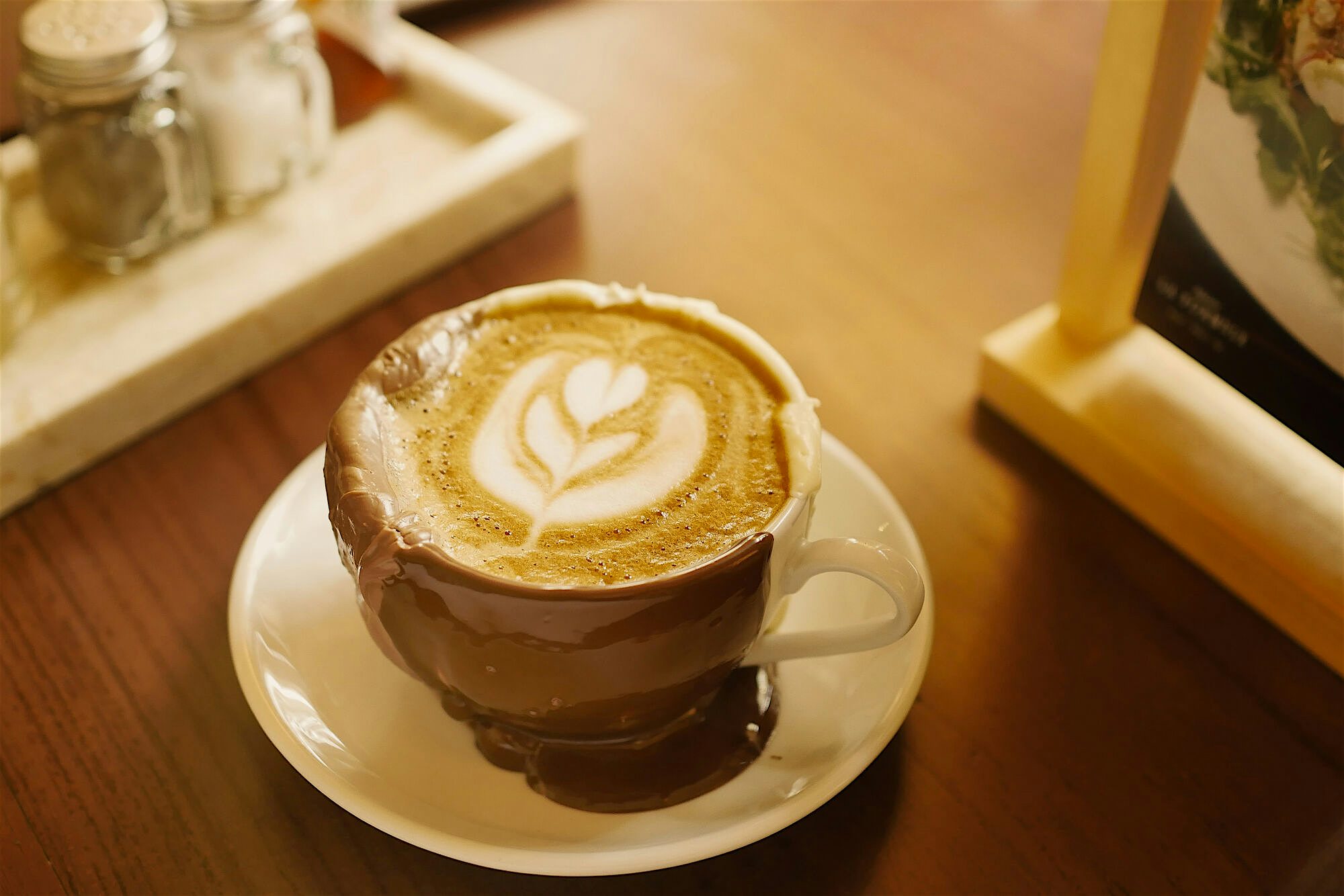 Cup of coffee with leaf-shaped latte art, served on a saucer with chocolate sauce dripped down the sides, placed on a wooden café table.
