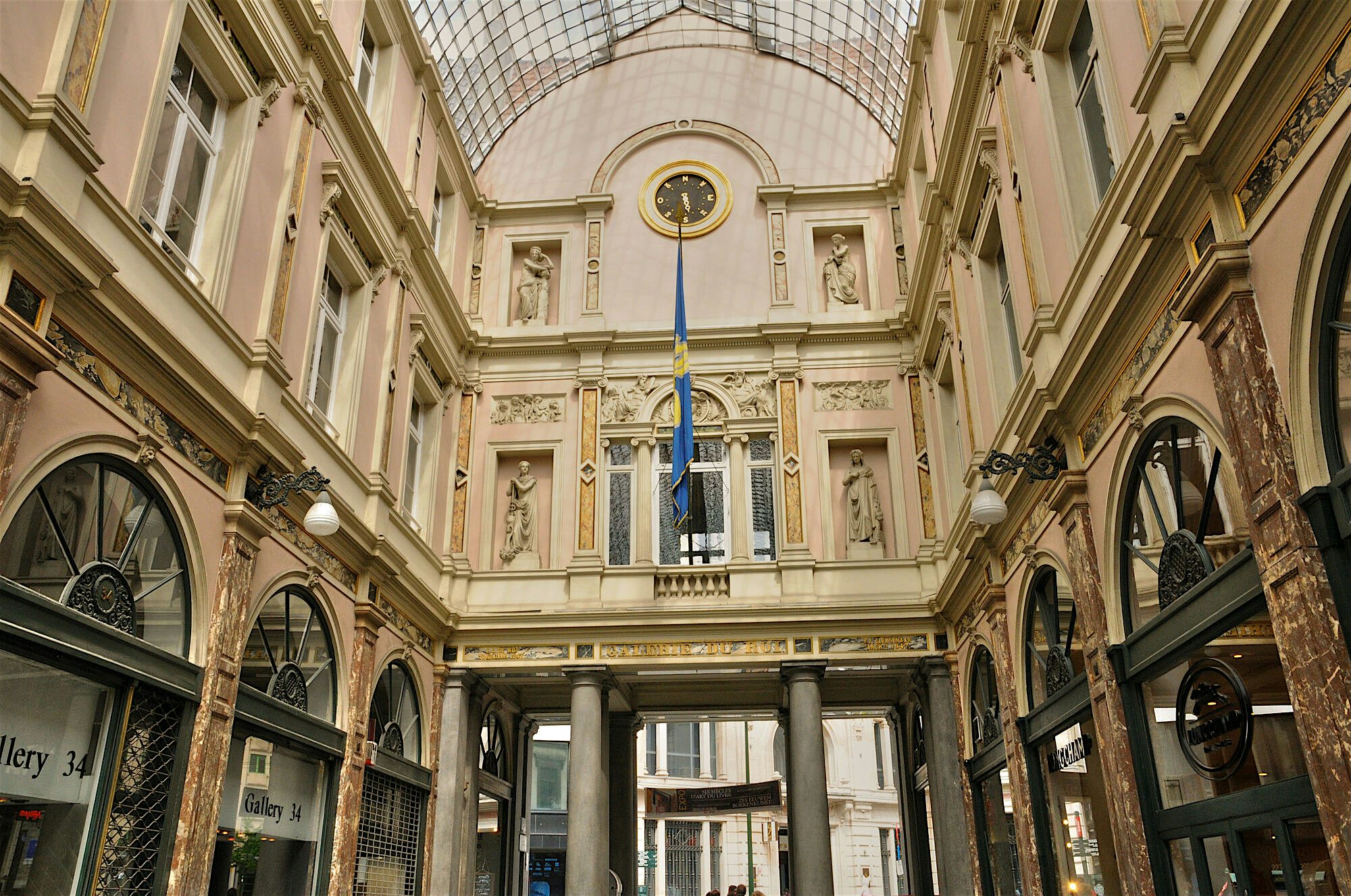 Ornate covered shopping arcade with carved stone details, statues, and a glass ceiling, framing a central passage beneath a clock and flag.