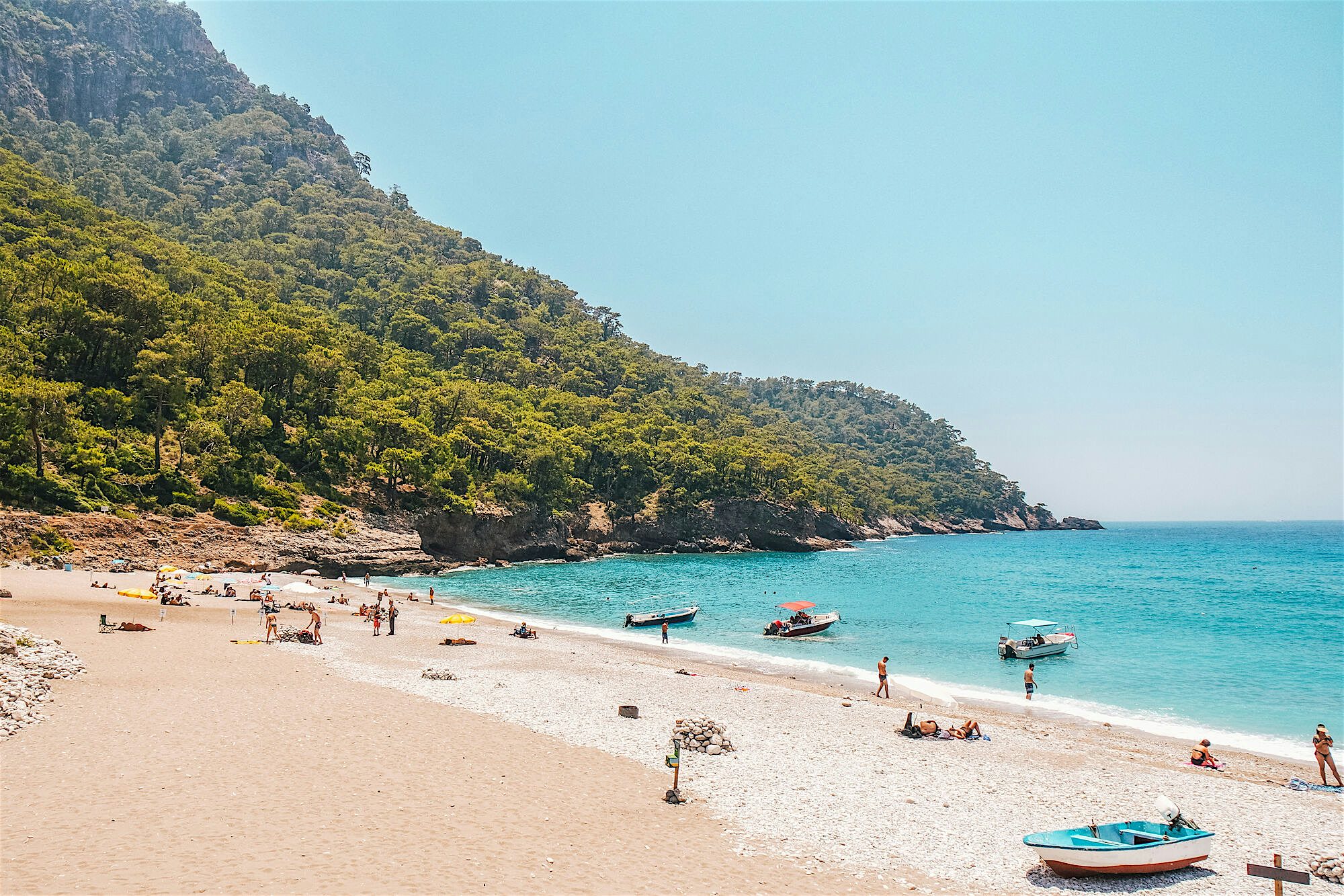 Pebbly beach, turquoise water, surrounding mountains covered in pine forests