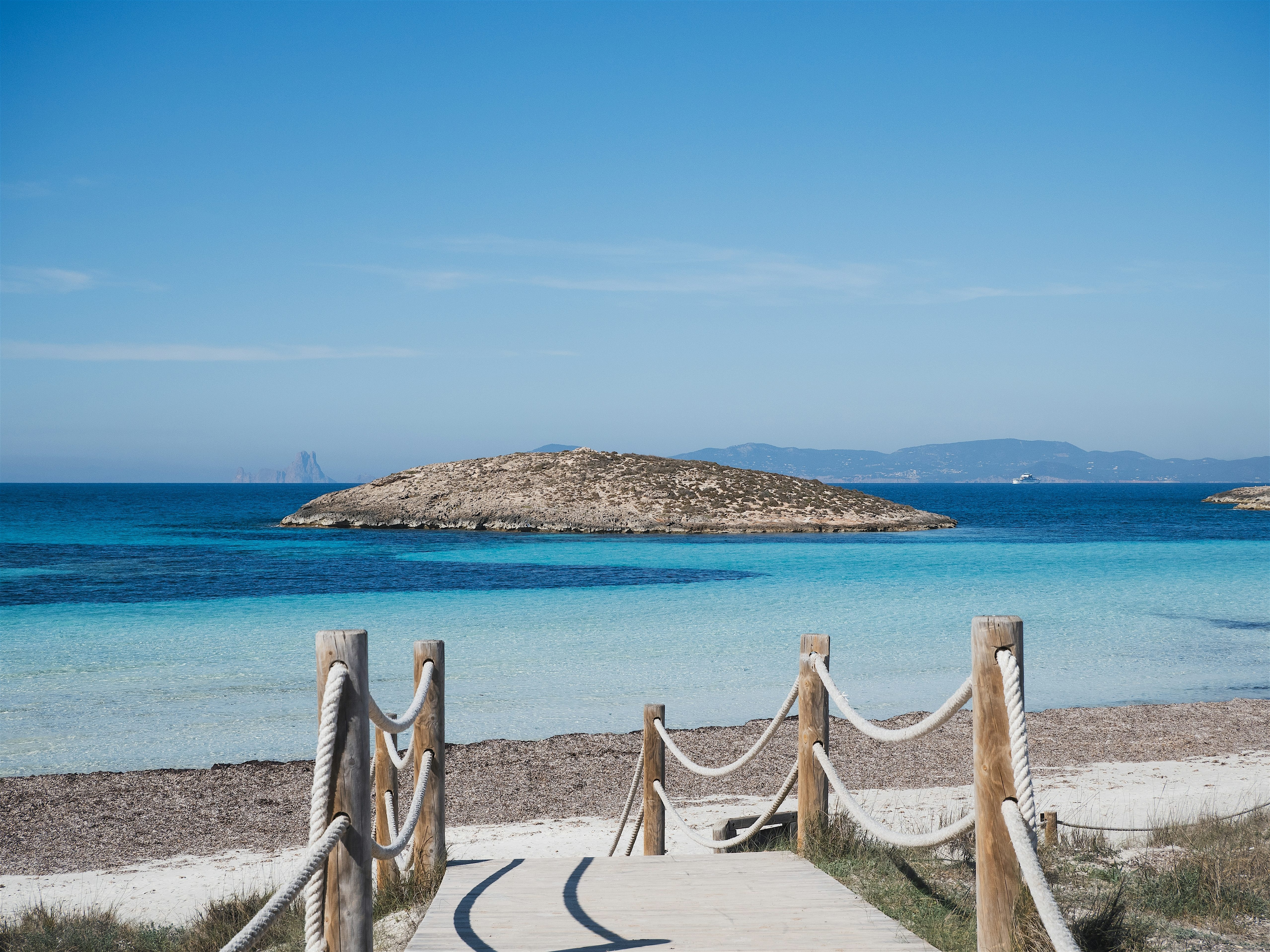 A wooden walkway leading down to a calm turquoise beach, facing a small rocky island just offshore.