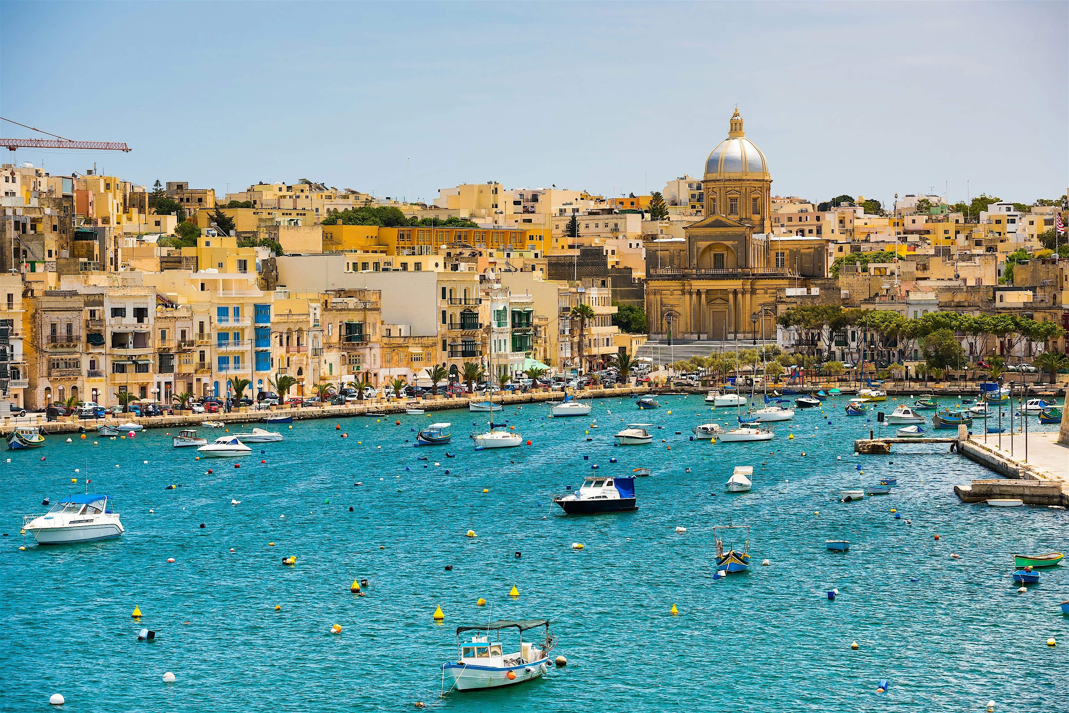 Colourful waterfront town beside a sheltered harbour, with small boats floating on bright blue water and a large domed church rising among tightly packed buildings along the shoreline.