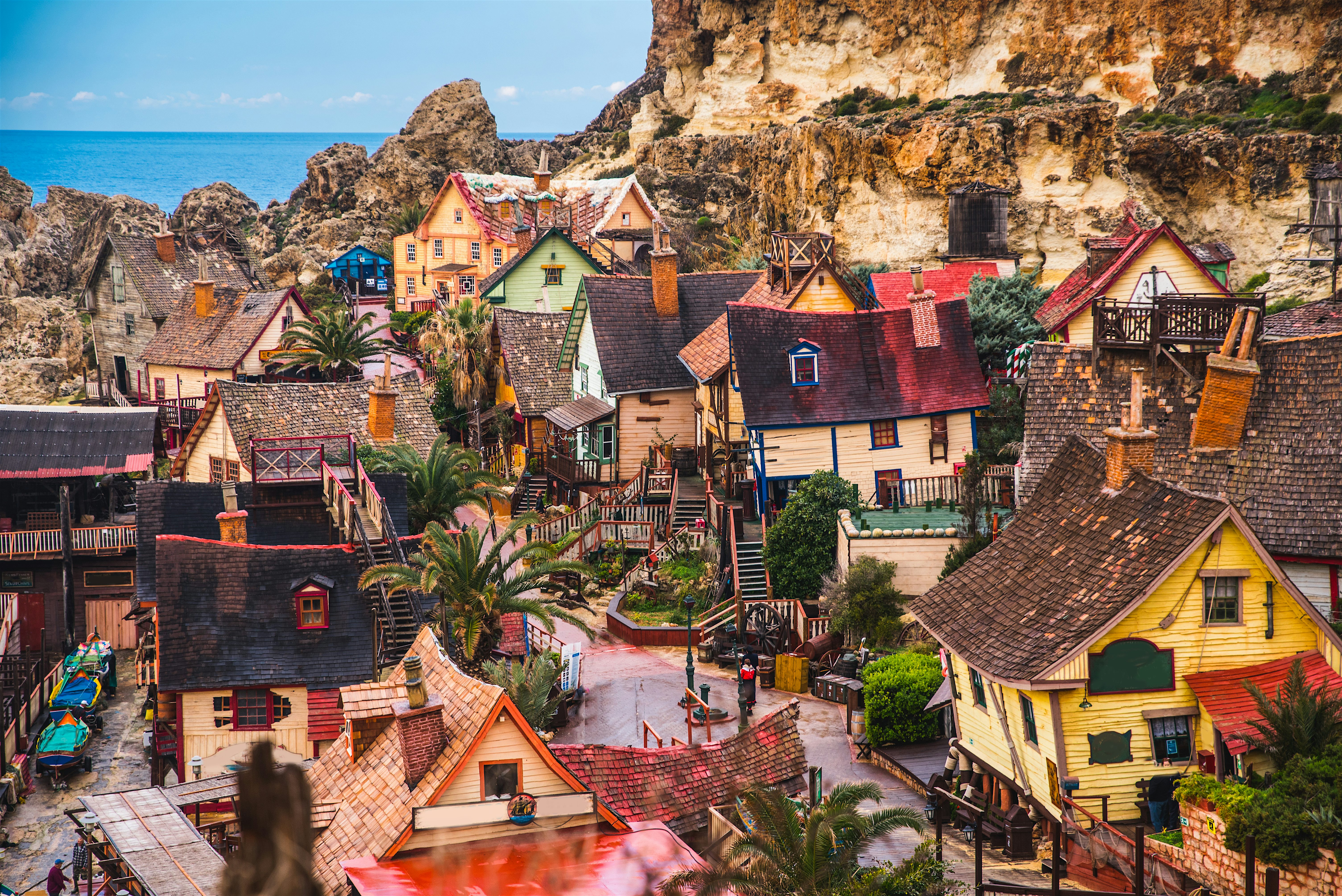 Cluster of brightly coloured cottages with nestled against rocky cliffs, featuring winding paths, terraces, and palm trees, with glimpses of blue sea visible beyond the village.