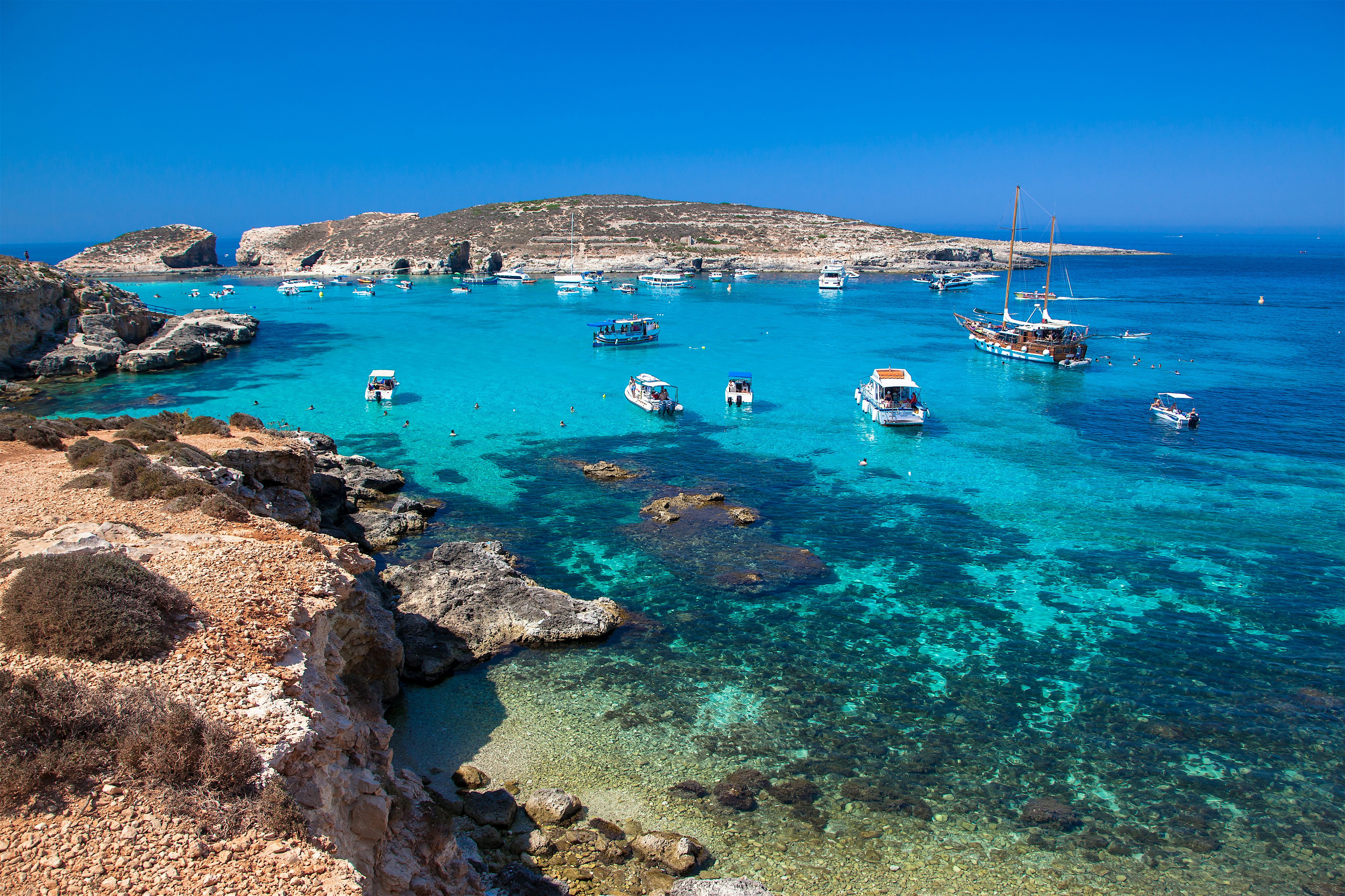 Clear turquoise bay with rocky shoreline in the foreground, dotted with small motorboats and yachts, and a low rugged island.