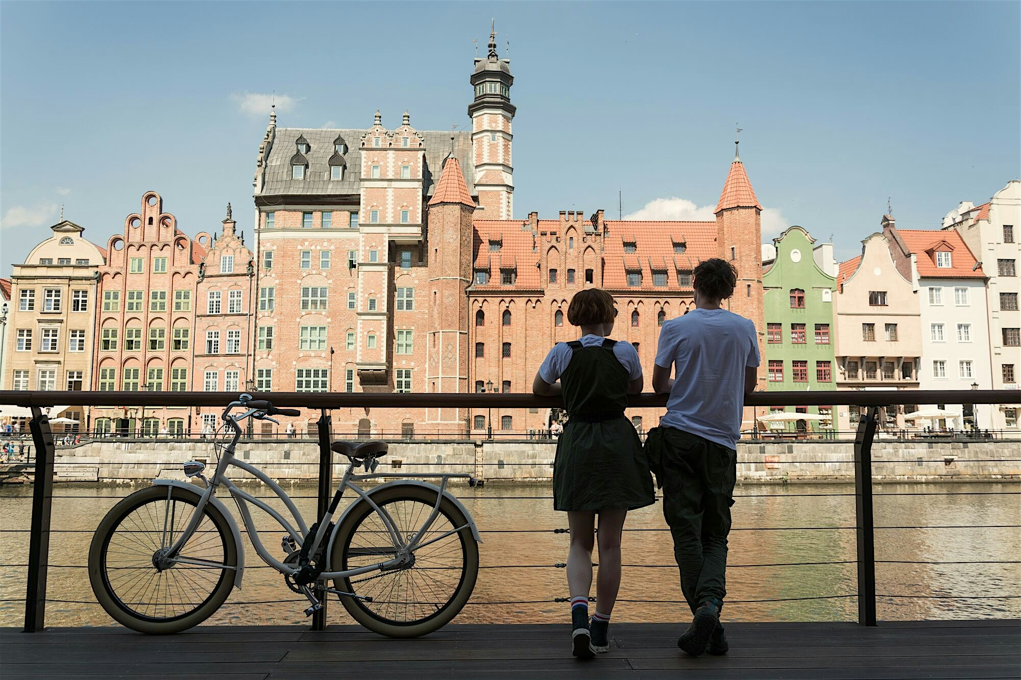 Two people leaning against a riverside railing with a bicycle beside them, looking out across water toward colourful historic buildings under a clear sky.
