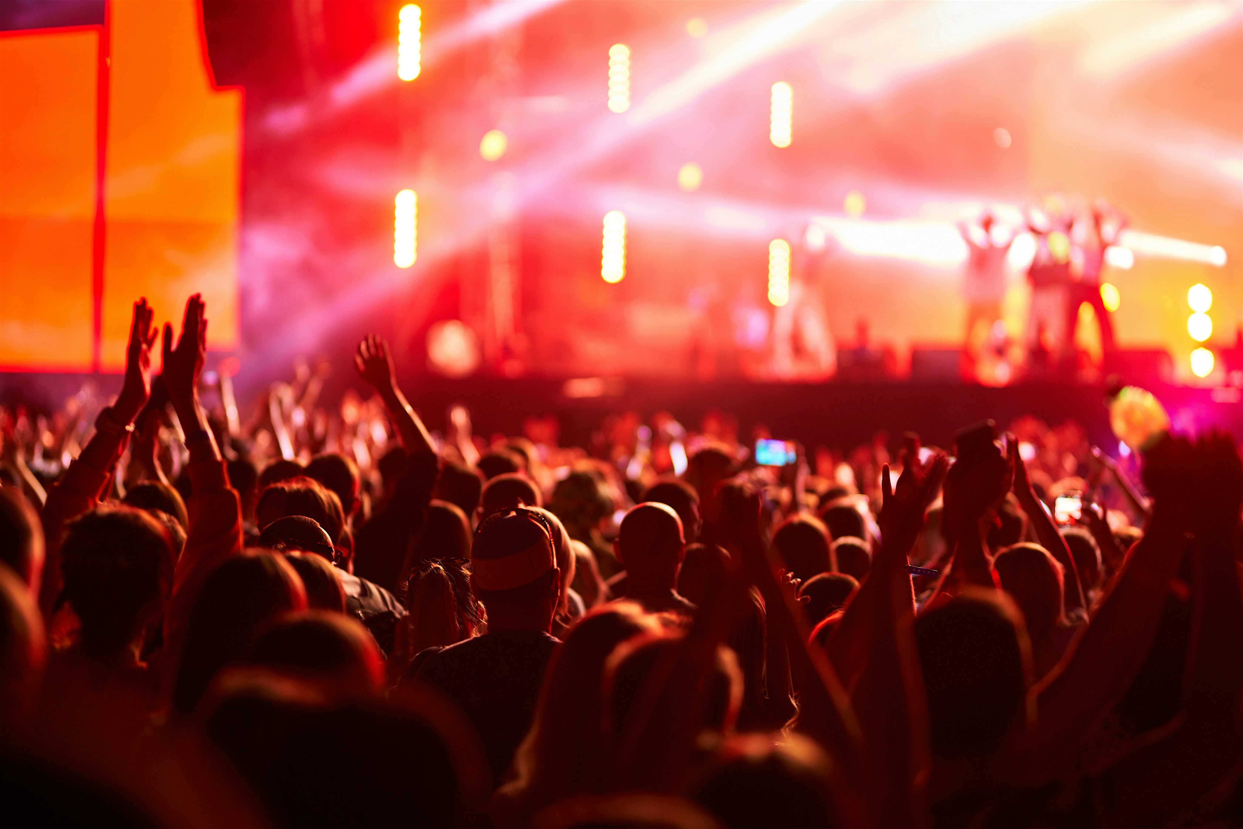 View from inside a music festival looking onto the stage. Pink and orange lights fill the area, with a range of bright strobe lighting