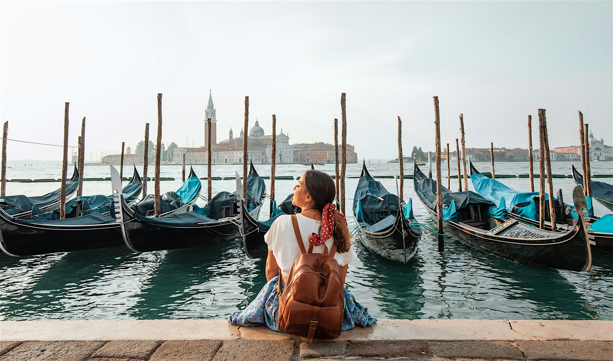 A person sitting by the waterfront looking out at a row of gondolas moored in the water, with historic buildings visible across the lagoon.