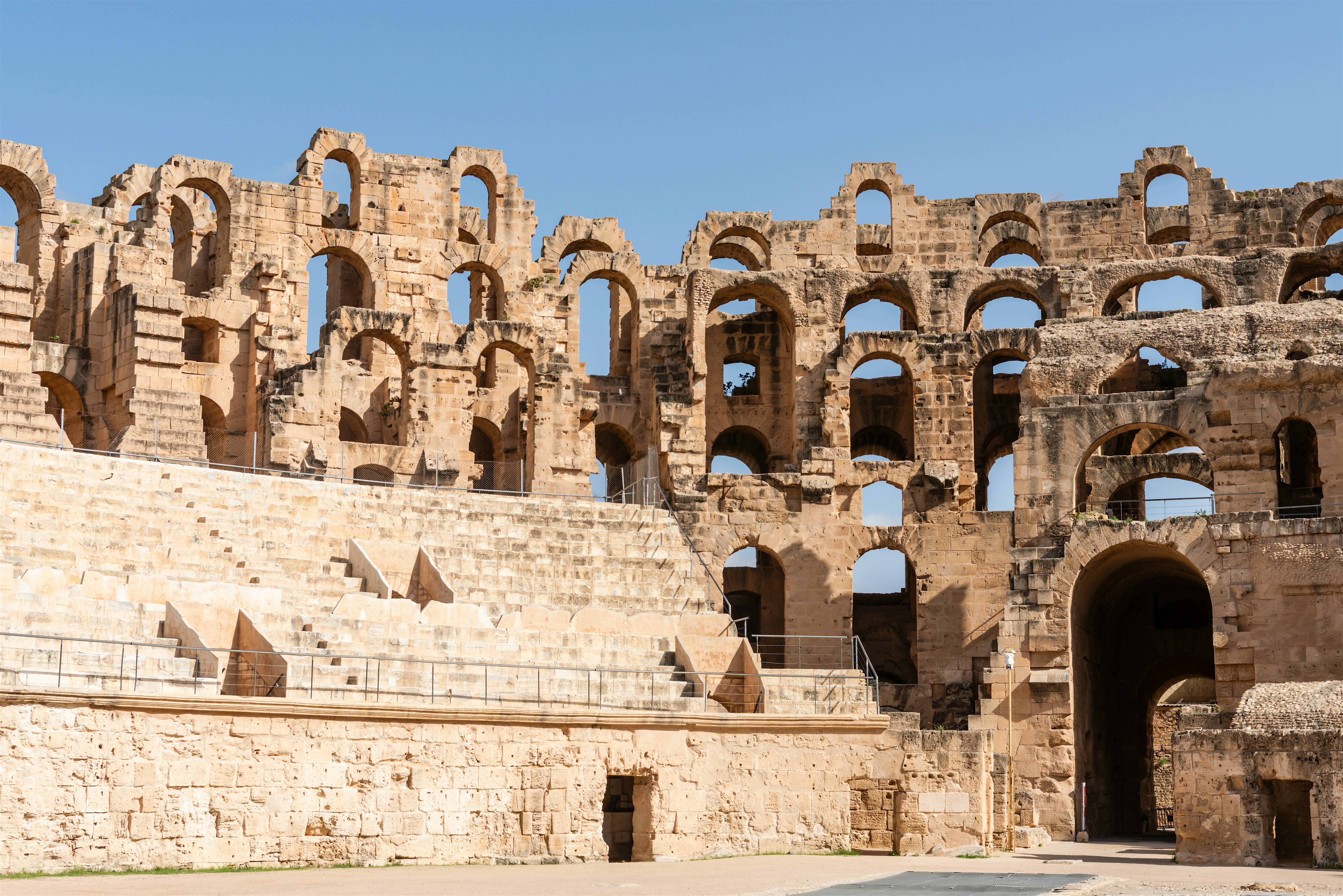 A large Roman amphitheatre with multiple stone arches and tiered seating rising above an open central arena