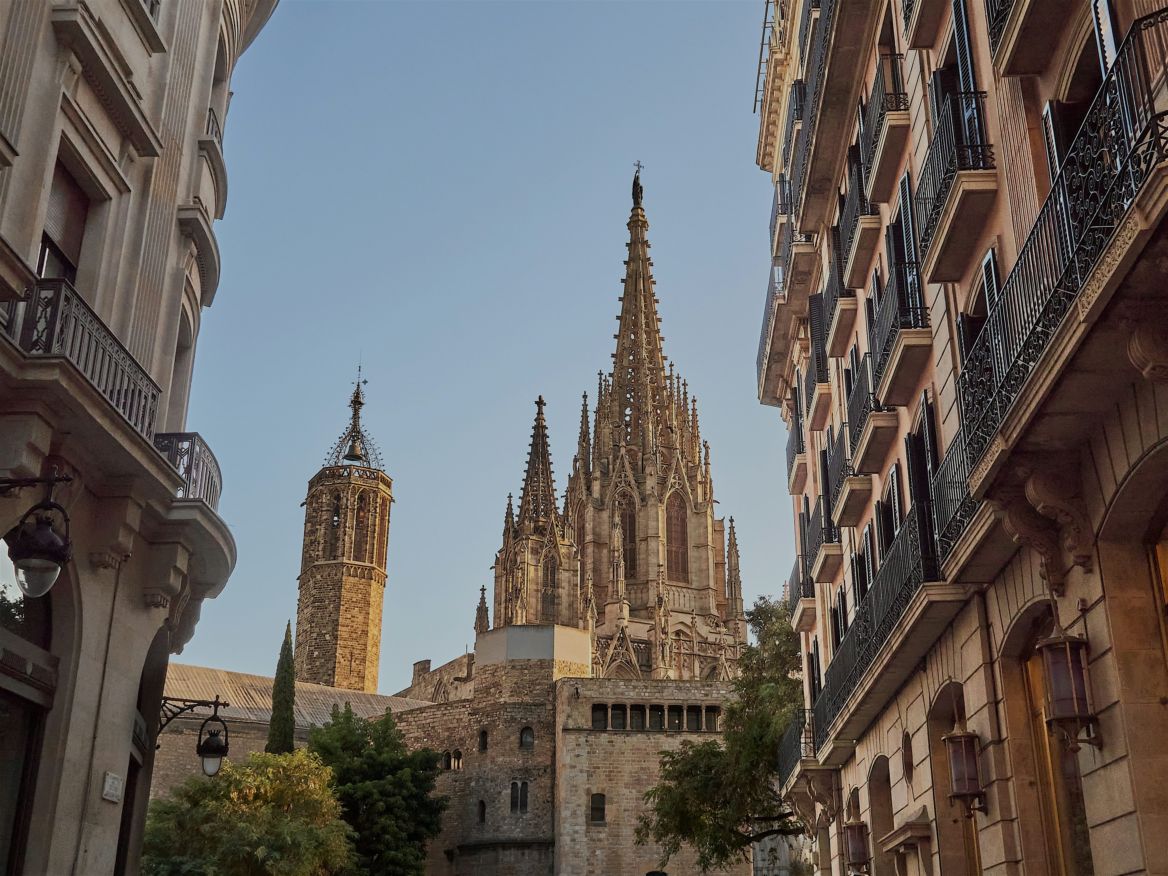 A bottom up view from a street, tradtitionally designed Spanish buildings. From in between the top of the Sagrada Familia can be seen with a clear blue sky behind