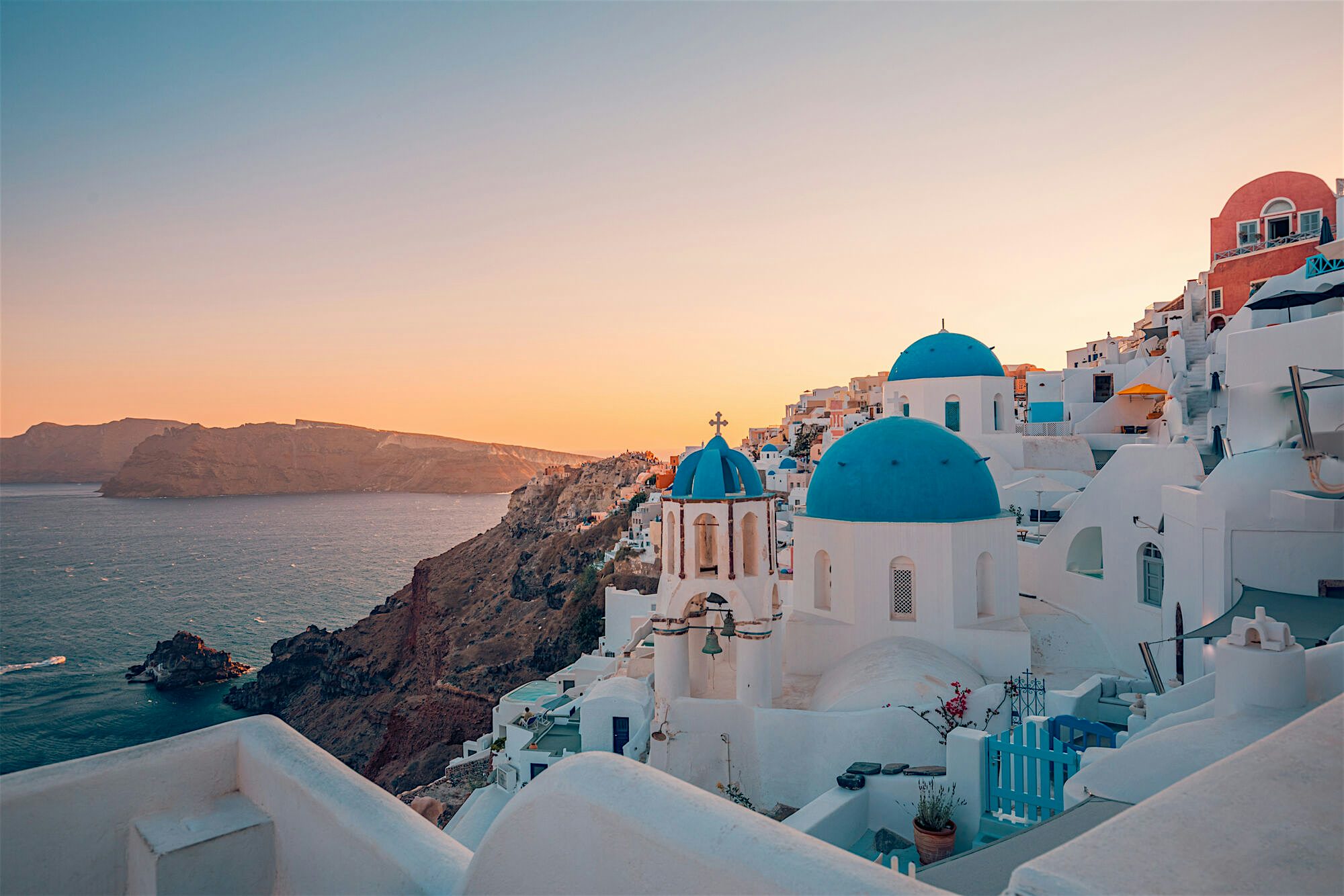 Whitewashed cliffside village at sunset, with blue‑domed churches and tightly packed buildings cascading down steep volcanic rock towards the sea, overlooking calm water